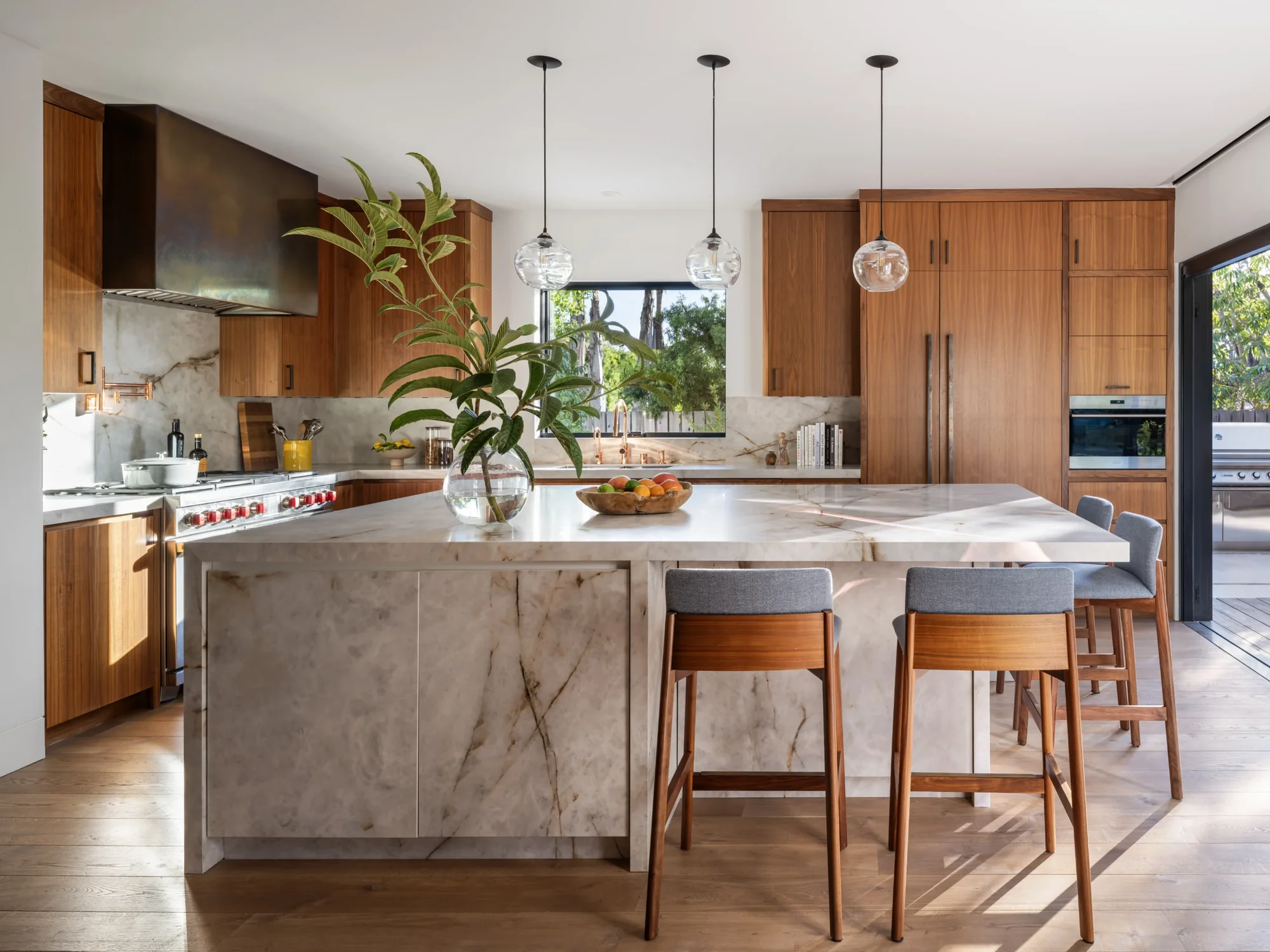 Modern kitchen with walnut cabinetry, marble island, glass pendant lights, and garden views through black-framed window.