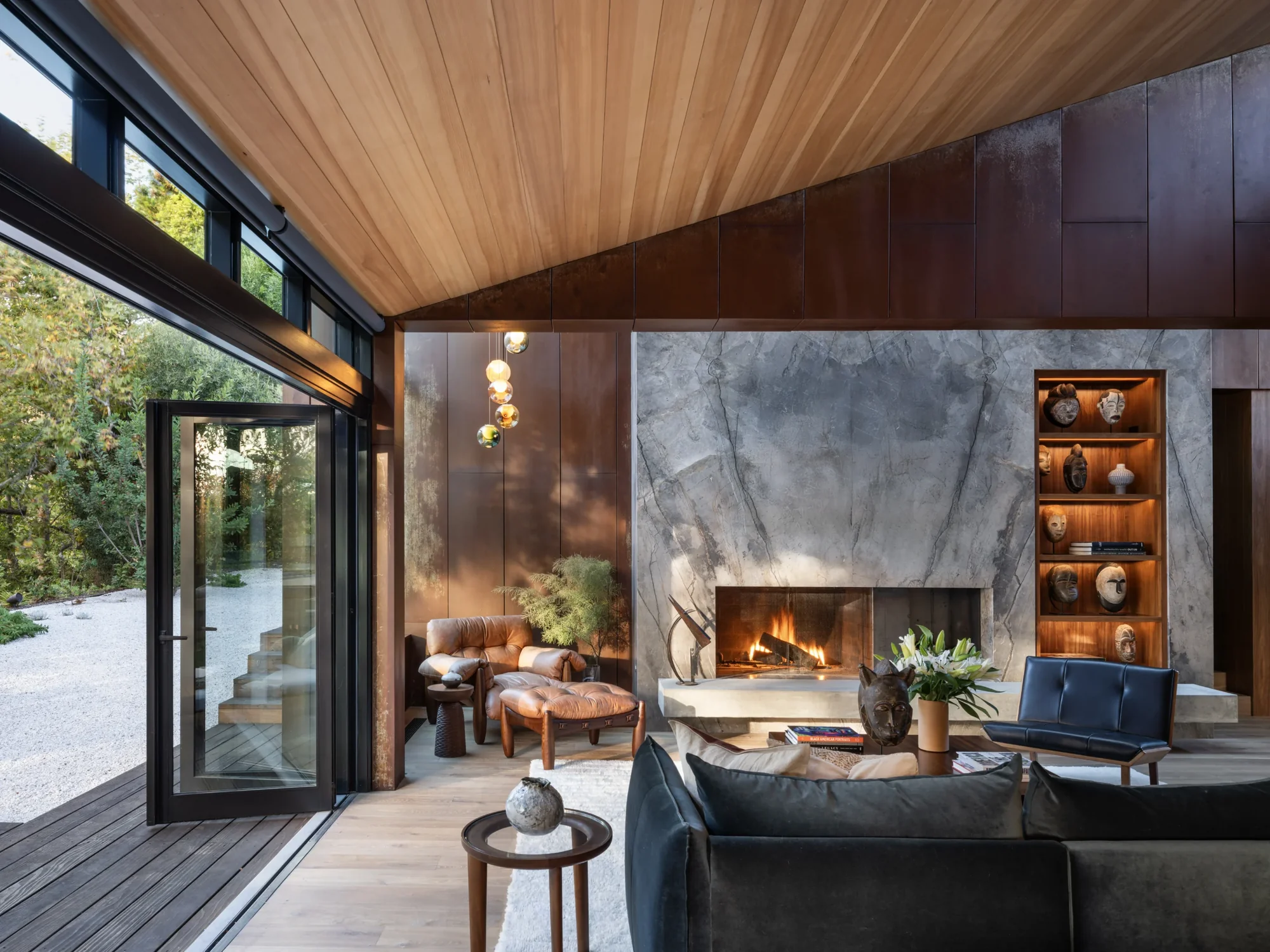 Modern living room with wood ceiling, marble fireplace wall, floor-to-ceiling windows, and leather seating opening to outdoor deck.