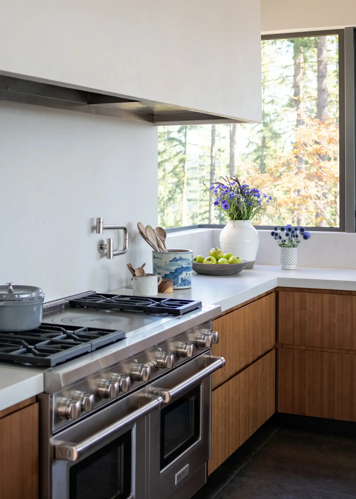 Modern kitchen with stainless gas range, wood cabinets, and large window framing forest views