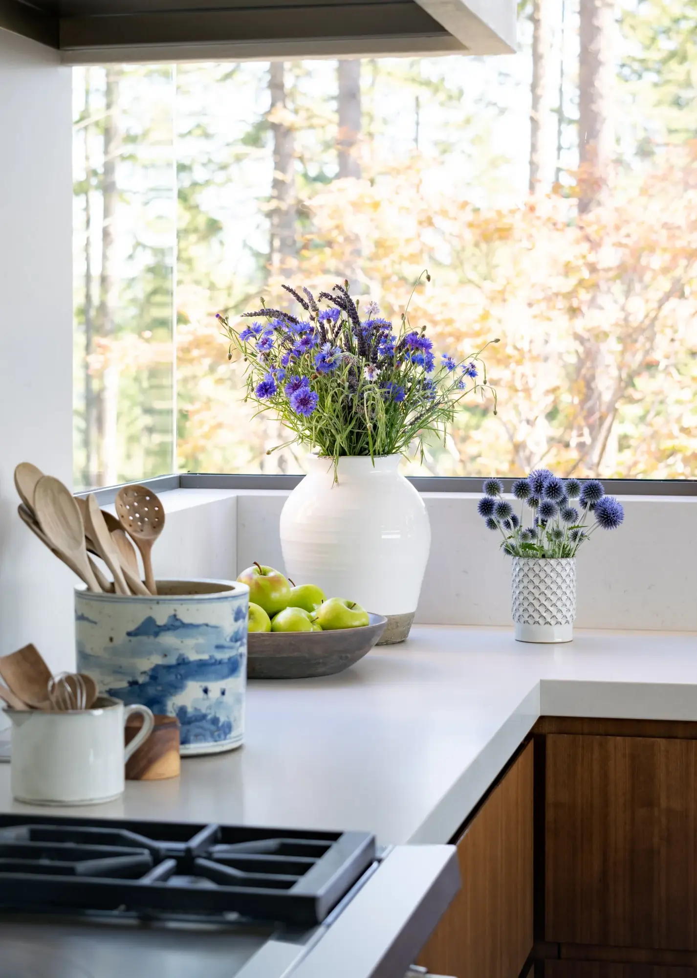Kitchen counter with purple wildflowers in white vases, wooden utensils, and green apples, overlooking autumn trees through large window