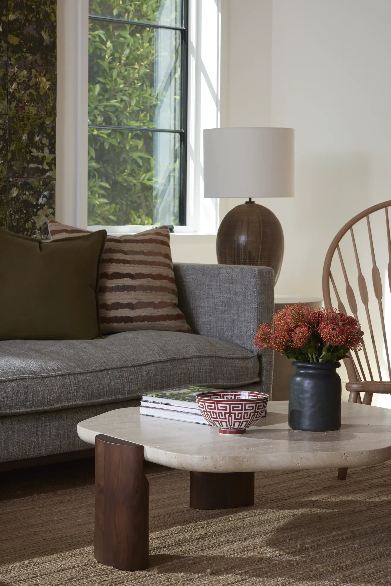 Gray sofa with striped pillows beside round stone coffee table and wooden lamp, large window showing greenery outside