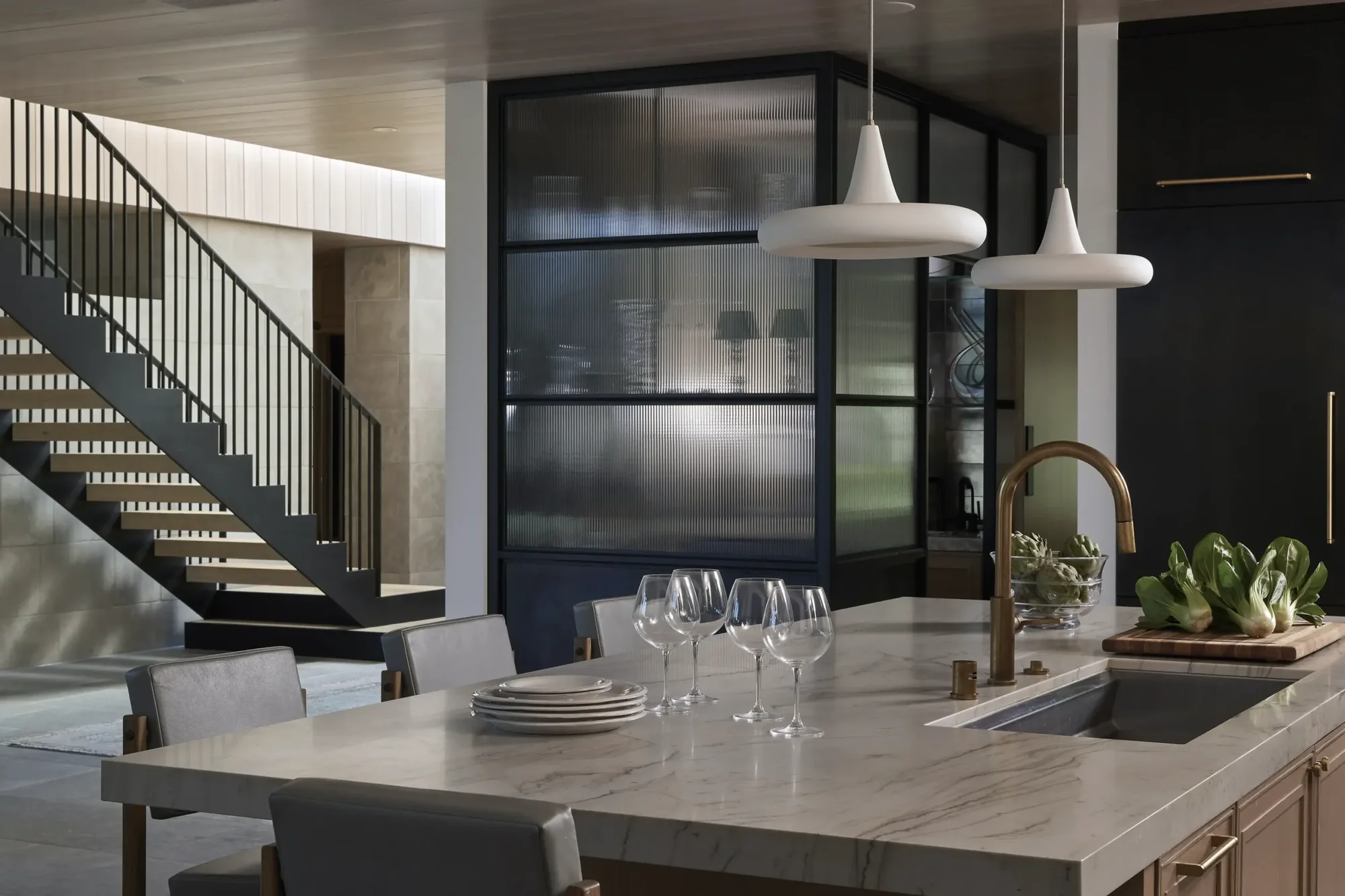 Modern kitchen island with marble countertop, wine glasses, and pendant lights, with black-framed glass partition and staircase beyond