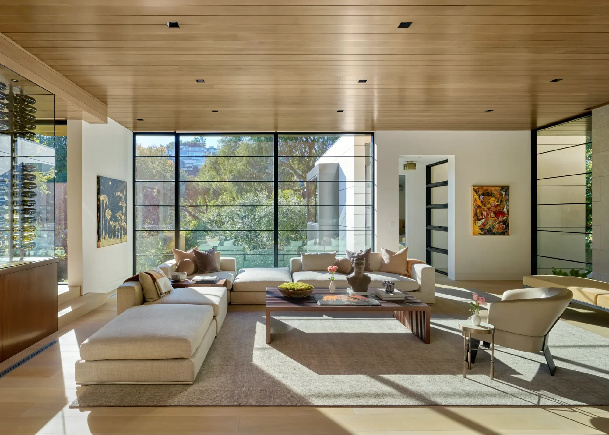Modern living room with wood ceiling, floor-to-ceiling windows overlooking garden, and neutral-toned sectional seating