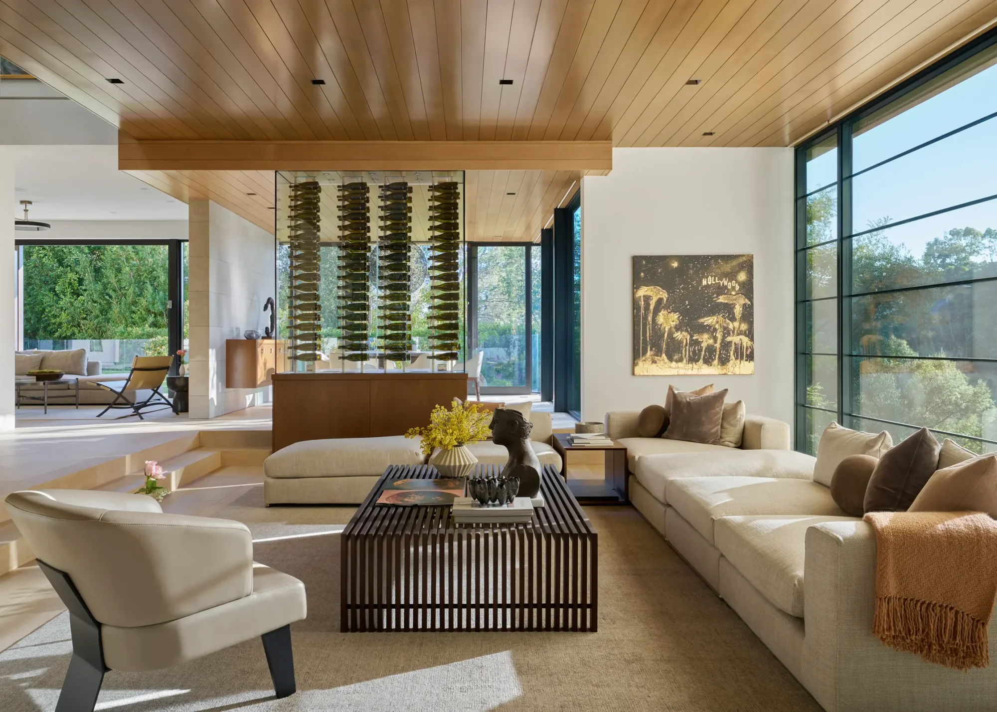 Modern living room with wood-planked ceiling, glass wine display, cream sofas, and floor-to-ceiling windows overlooking nature.