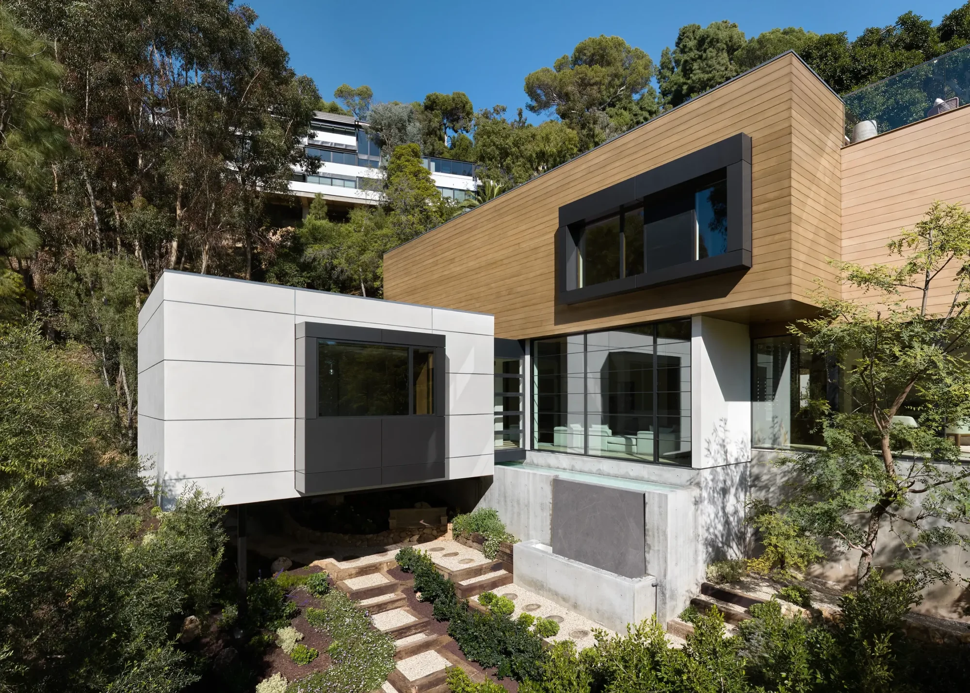 Modern hillside home with cedar upper level and white lower level, featuring large windows and landscaped steps.