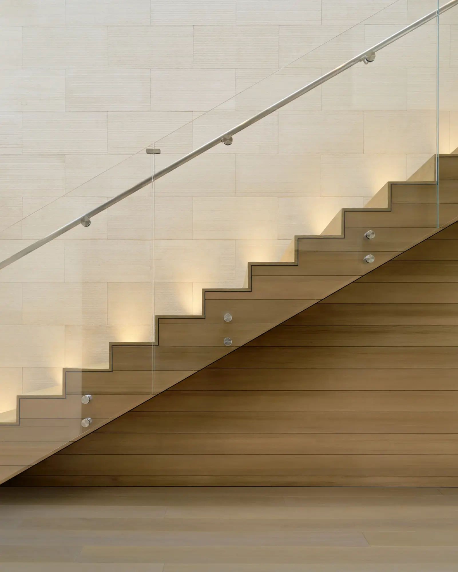 Modern wooden staircase with glass railing and metal handrail against textured white stone wall with integrated lighting.