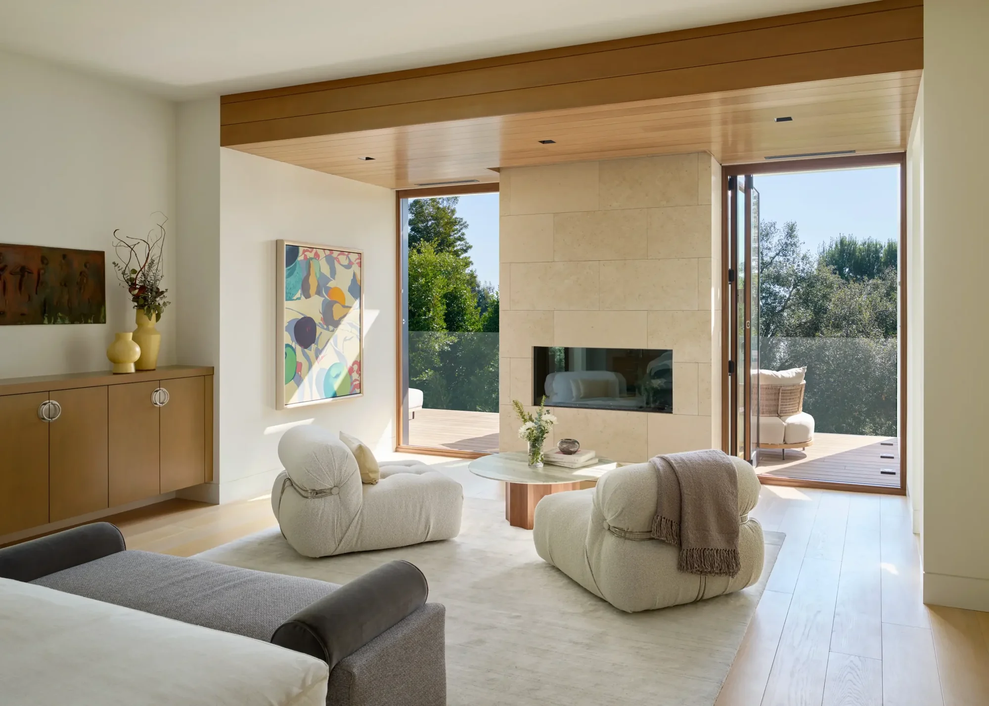 Modern living room with stone fireplace, cream lounge chairs, and open doors to outdoor terrace with wood ceiling