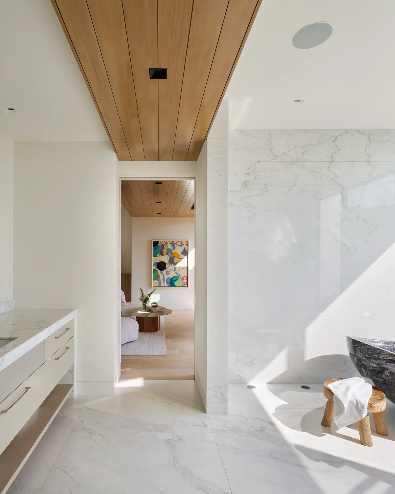 Modern bathroom with wood-paneled ceiling, white marble walls, and hallway view to living room with colorful artwork