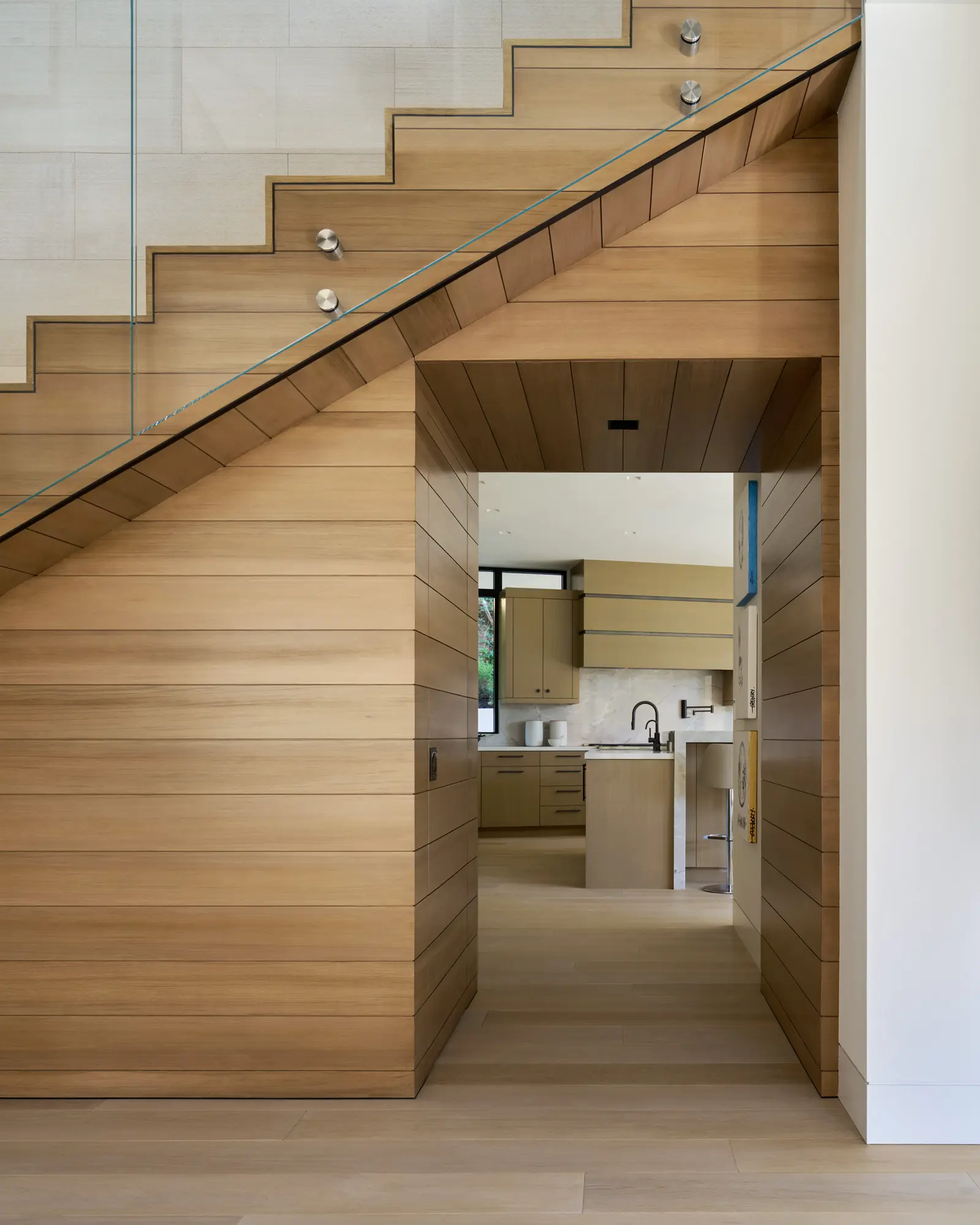 Wooden staircase with glass railing above kitchen entry featuring horizontal cedar paneling and natural materials