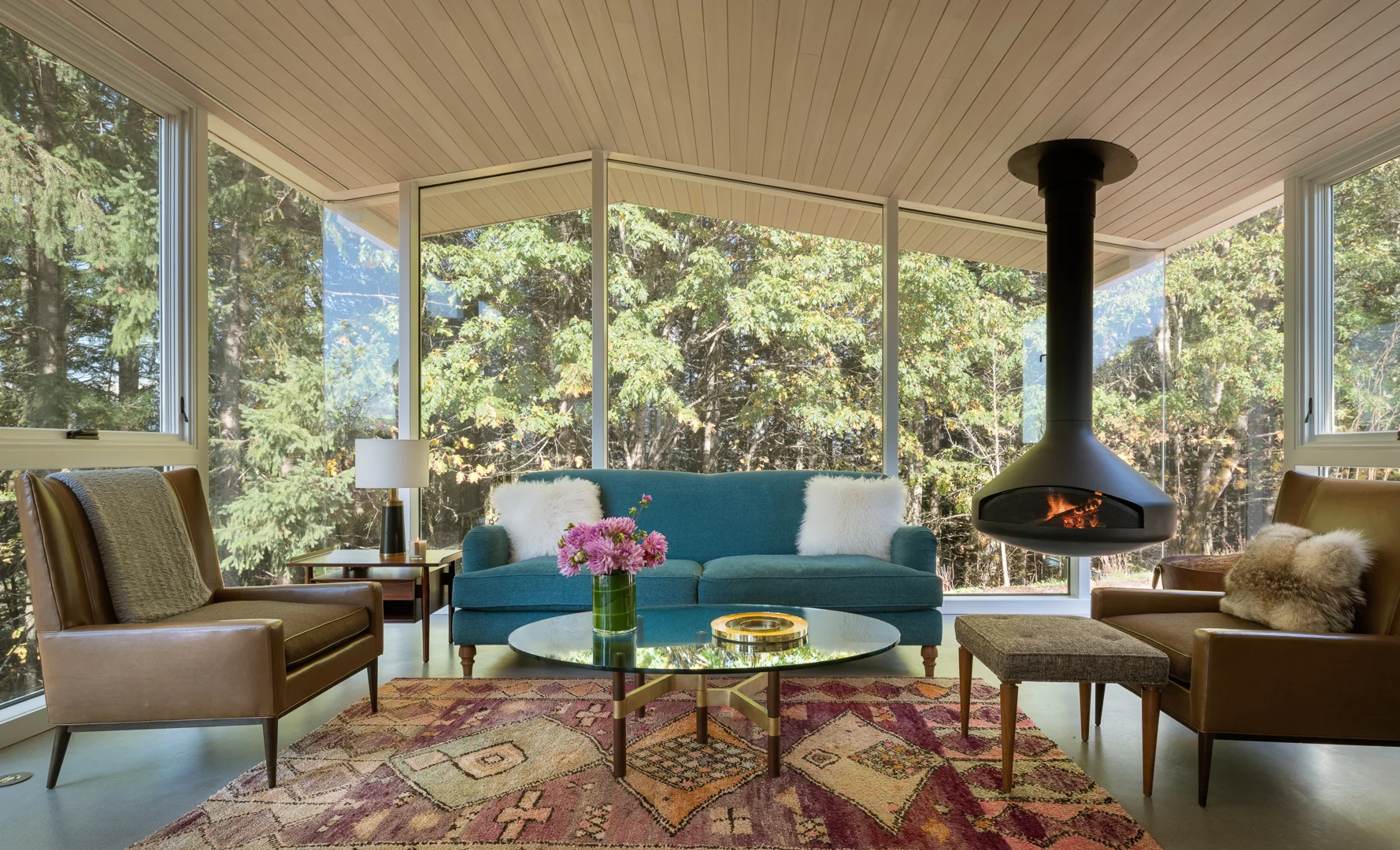 Mid-century modern living room with floor-to-ceiling windows, suspended fireplace, and mountain views through trees