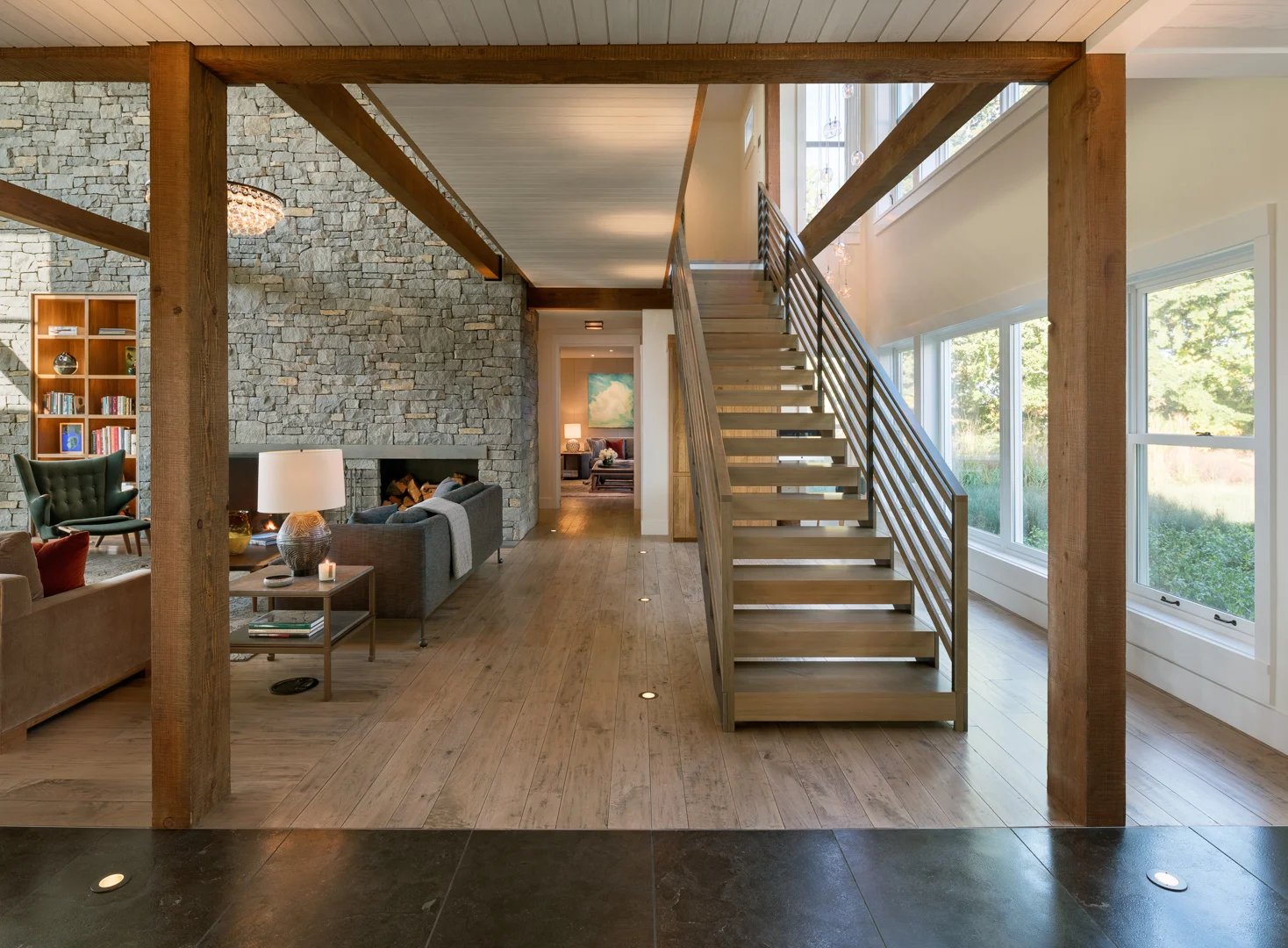 Open-concept living room with exposed timber beams, stone fireplace, floating wood stairs, and floor-to-ceiling windows.