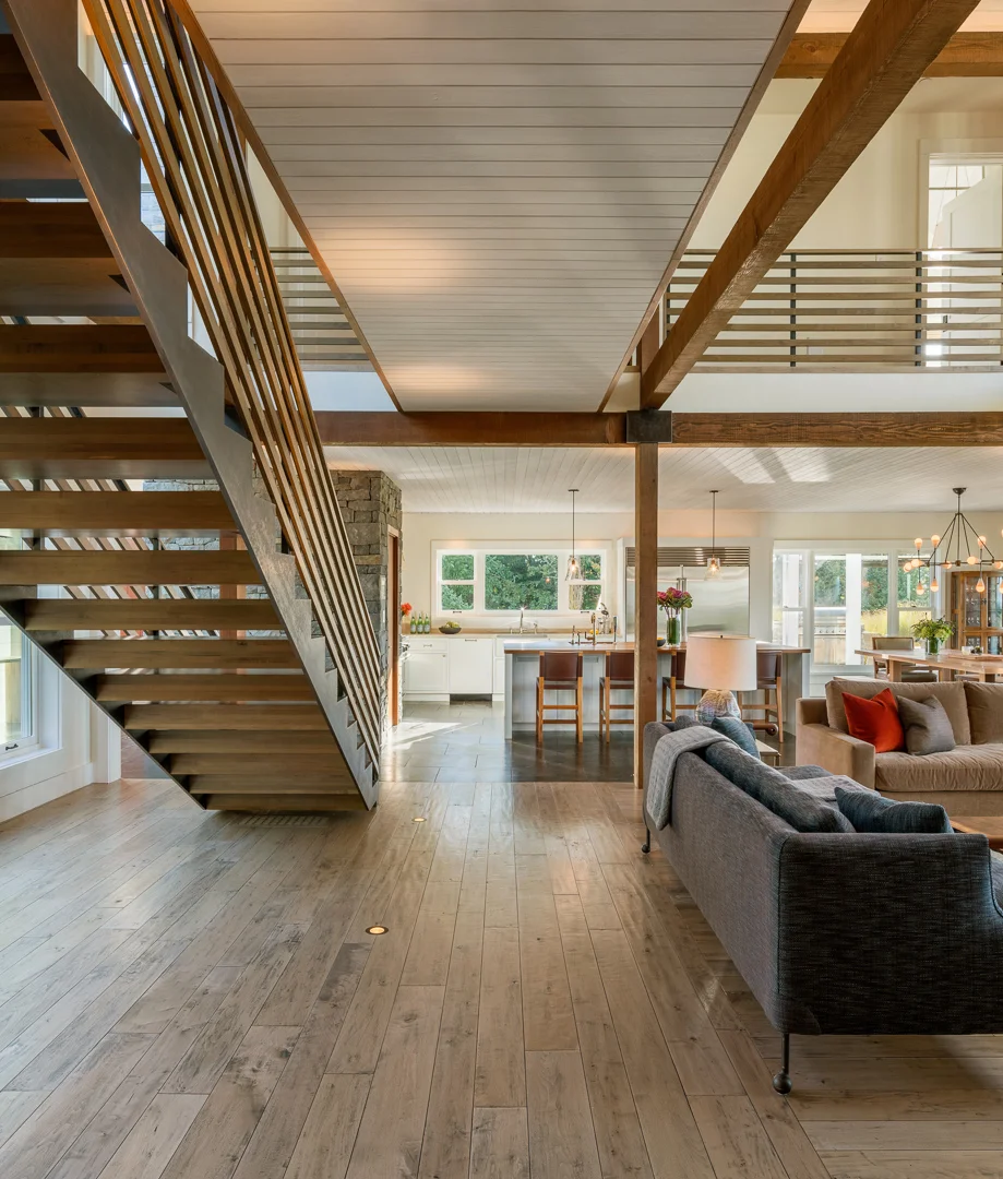 Open-concept living space with exposed timber beams, floating wooden staircase, and natural light flowing through kitchen