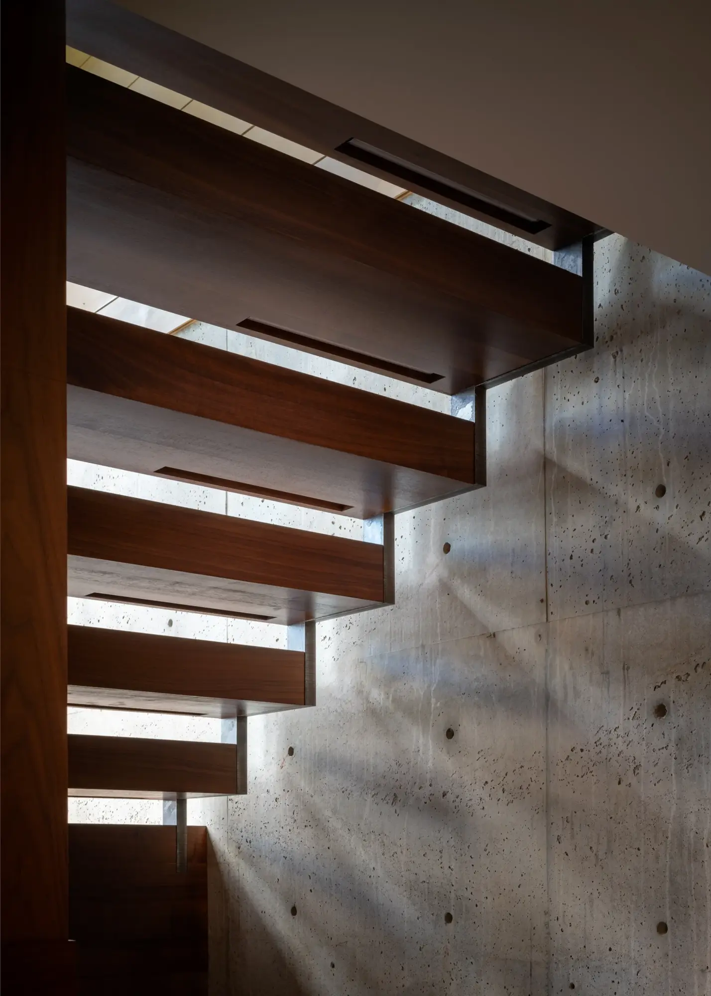 Floating wooden stairs against concrete wall with natural light filtering through horizontal slats
