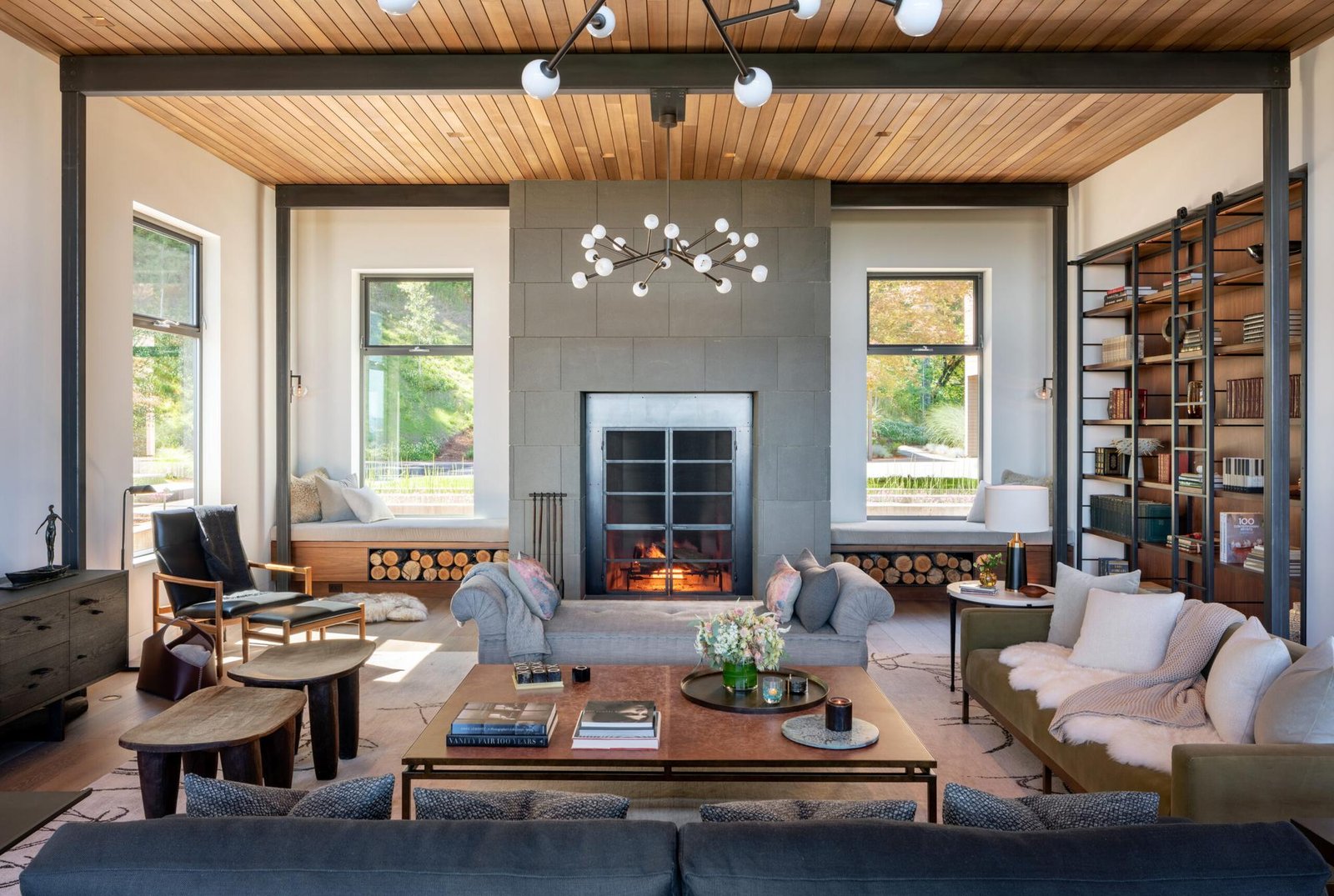 Modern living room with wood ceiling, stone fireplace, built-in window seats, and molecular chandelier in custom home