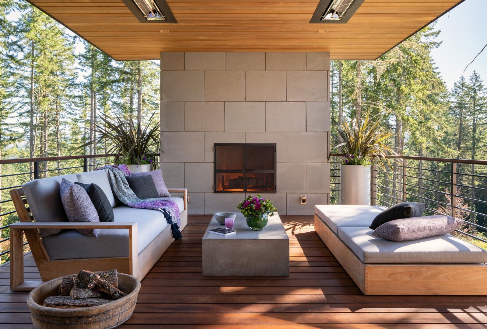 Covered outdoor deck with modern fireplace, wood furniture with gray cushions, and forest views beyond cable railing.