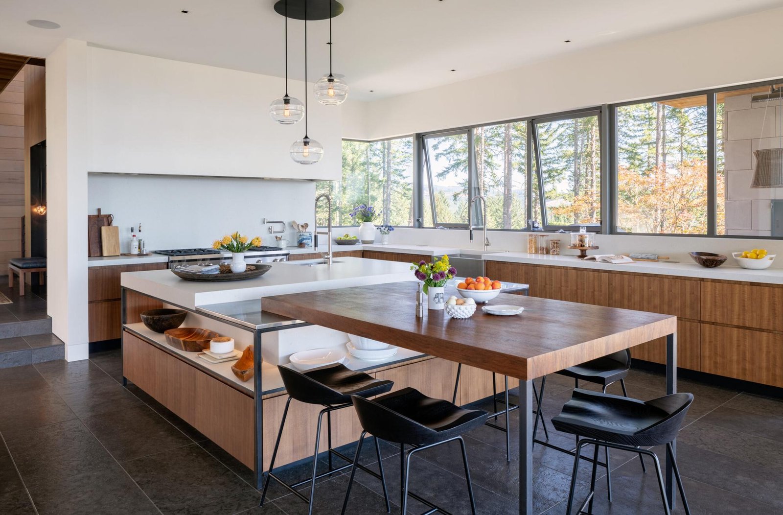 Modern kitchen with walnut cabinetry, white countertops, island dining table, and large windows overlooking forest