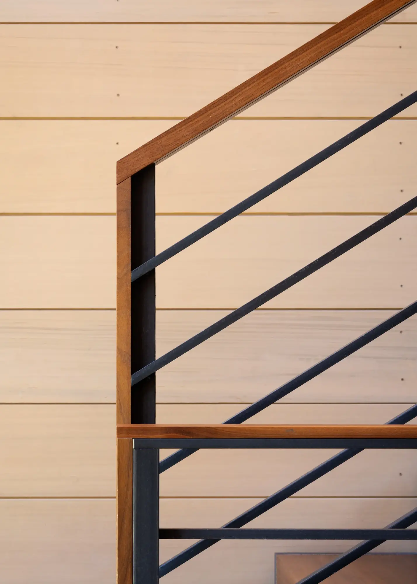 Modern stair railing with wood handrail and black metal diagonal bars against light wood siding.