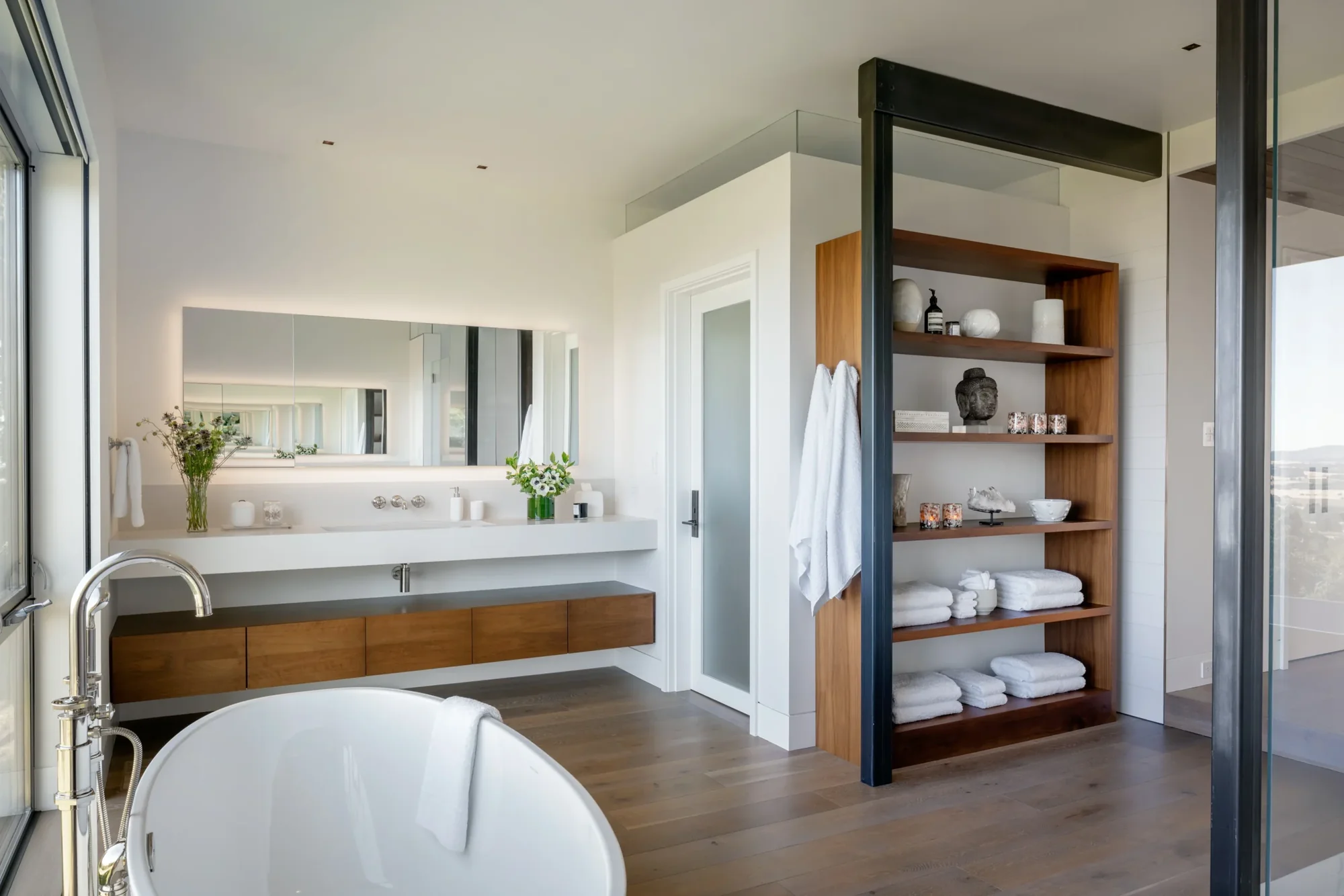 Modern bathroom with freestanding tub, floating wood vanity, built-in shelving, and wood flooring with natural light