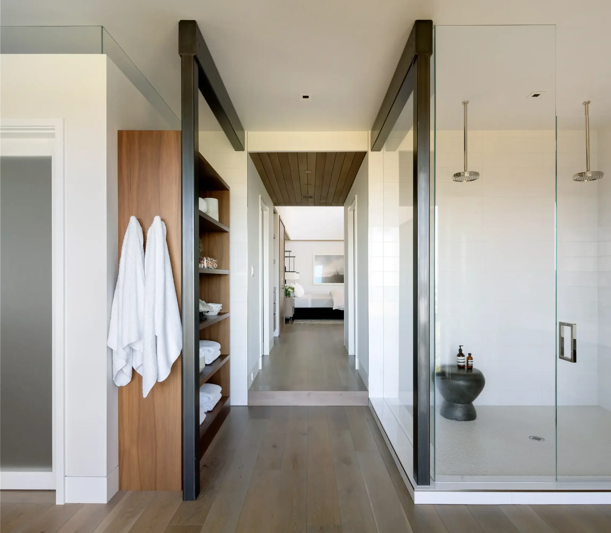 Modern bathroom with glass-enclosed shower, wooden shelving with white towels, and hallway leading to bedroom beyond