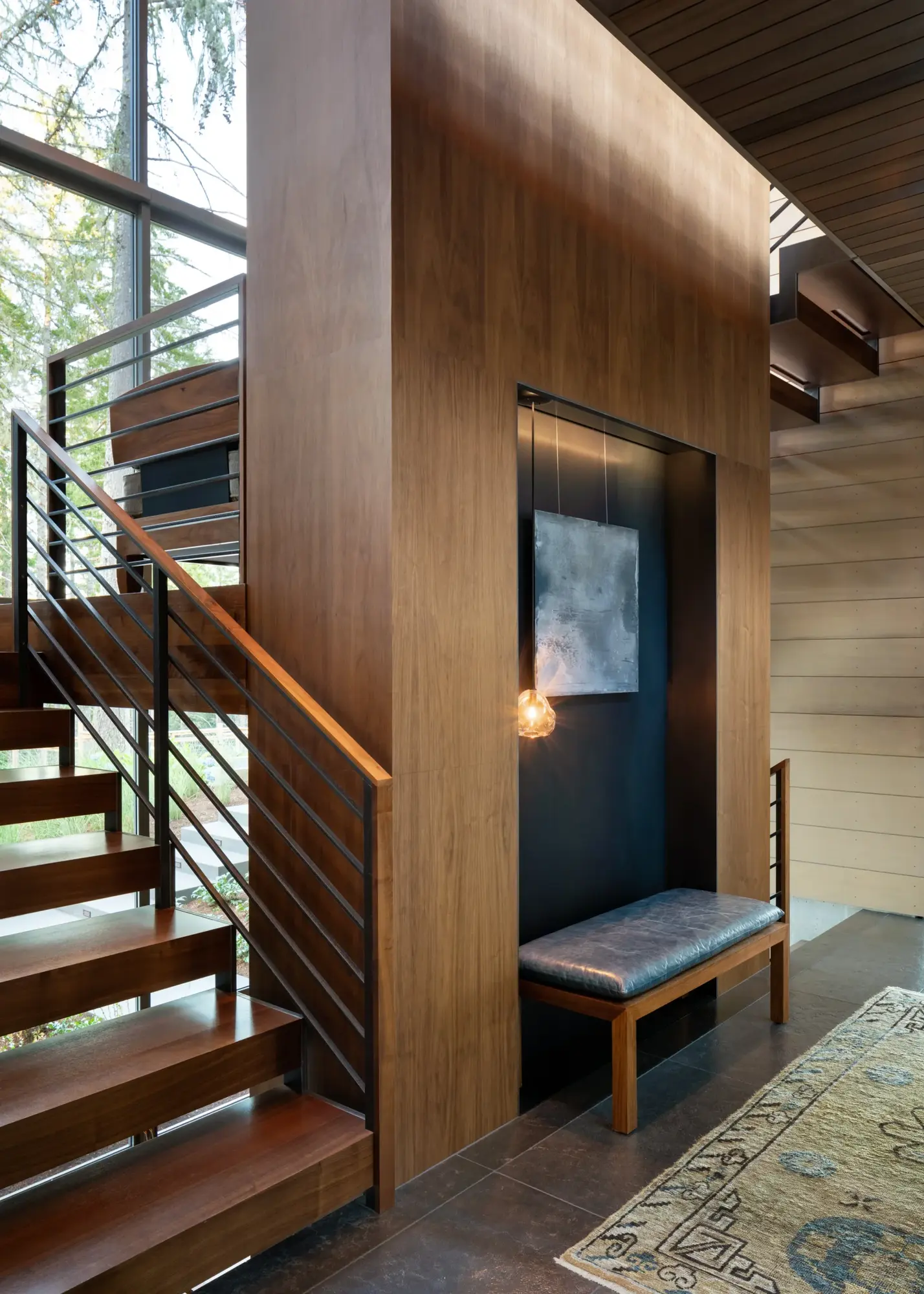 Modern wood-paneled entryway with open staircase, dark accent wall, bench seating, and natural light from tall windows.