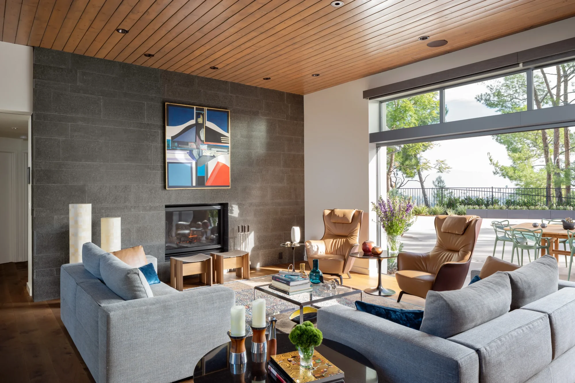 Modern living room with wood ceiling, gray stone fireplace, leather chairs, gray sofas, and floor-to-ceiling windows opening to outdoor patio.