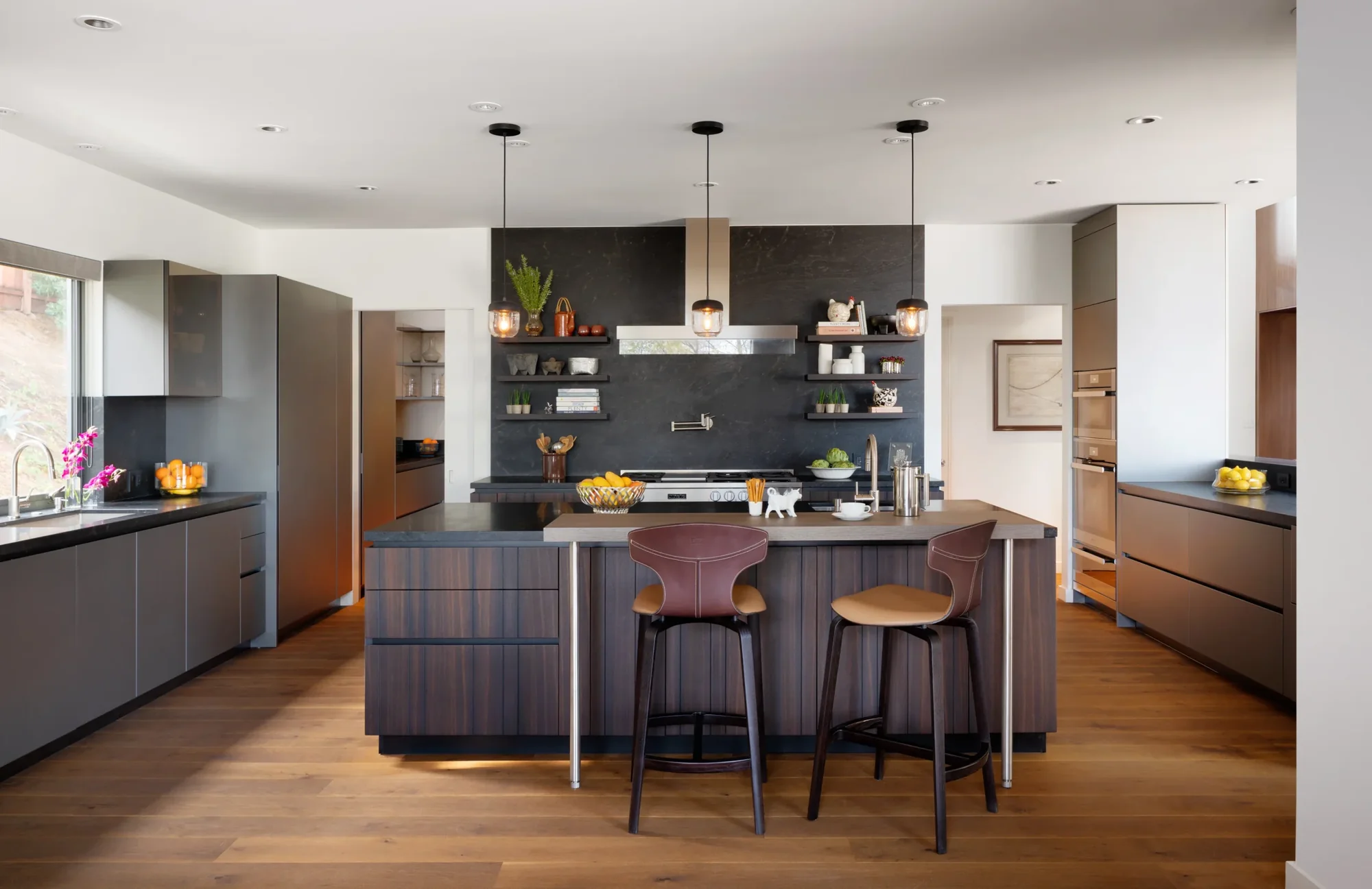 Modern kitchen with dark wood island, black accent wall with open shelving, and pendant lighting over natural wood floors.
