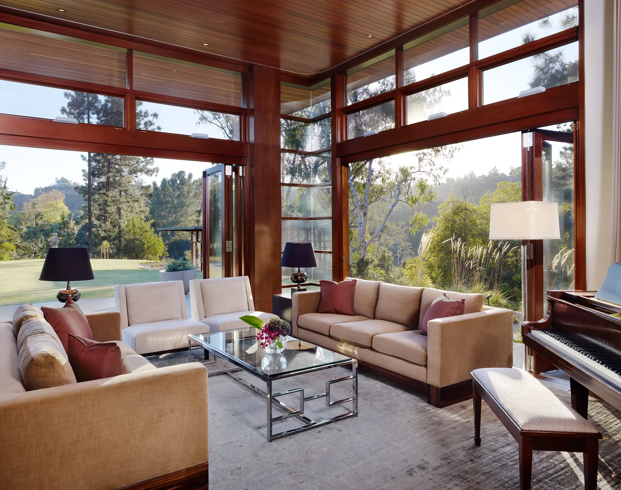 Modern living room with floor-to-ceiling windows, neutral sofas, and wood ceiling overlooking lush landscape