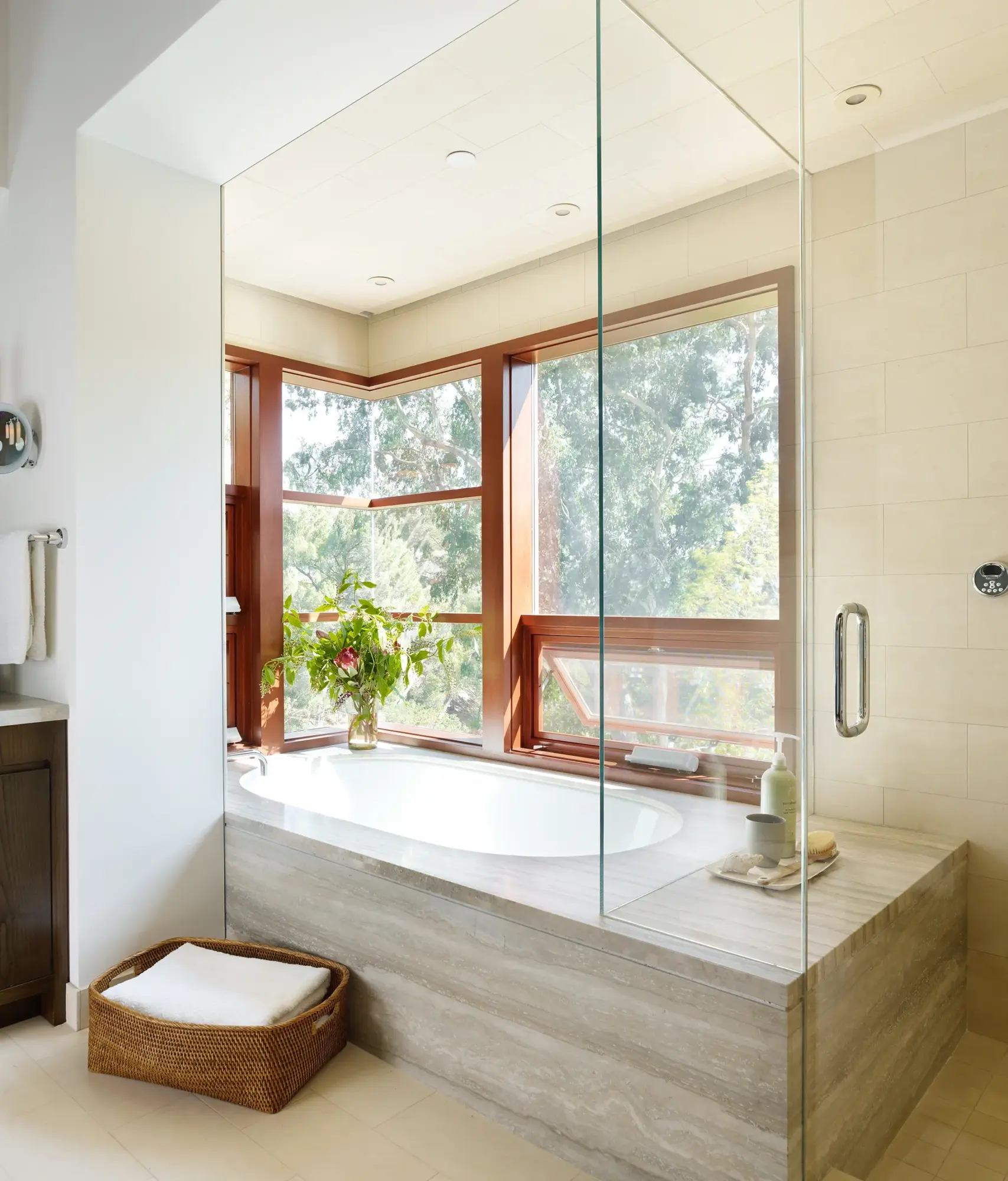 Modern bathroom with soaking tub framed by mahogany windows overlooking trees, glass shower partition, natural stone surround