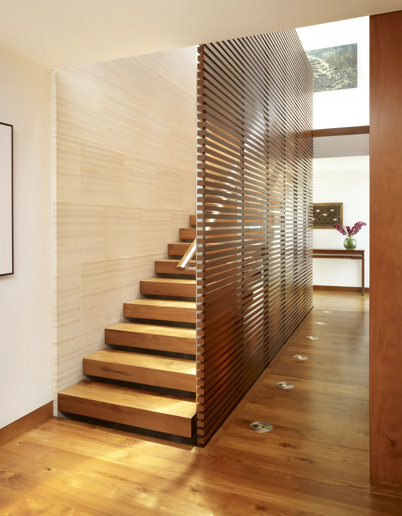 Floating wooden staircase with horizontal slat privacy screen, floor lighting, and travertine accent wall