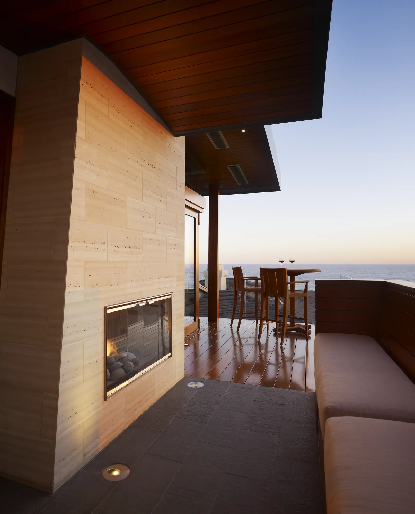 Modern outdoor patio with stone fireplace, wooden dining area, and ocean views at twilight