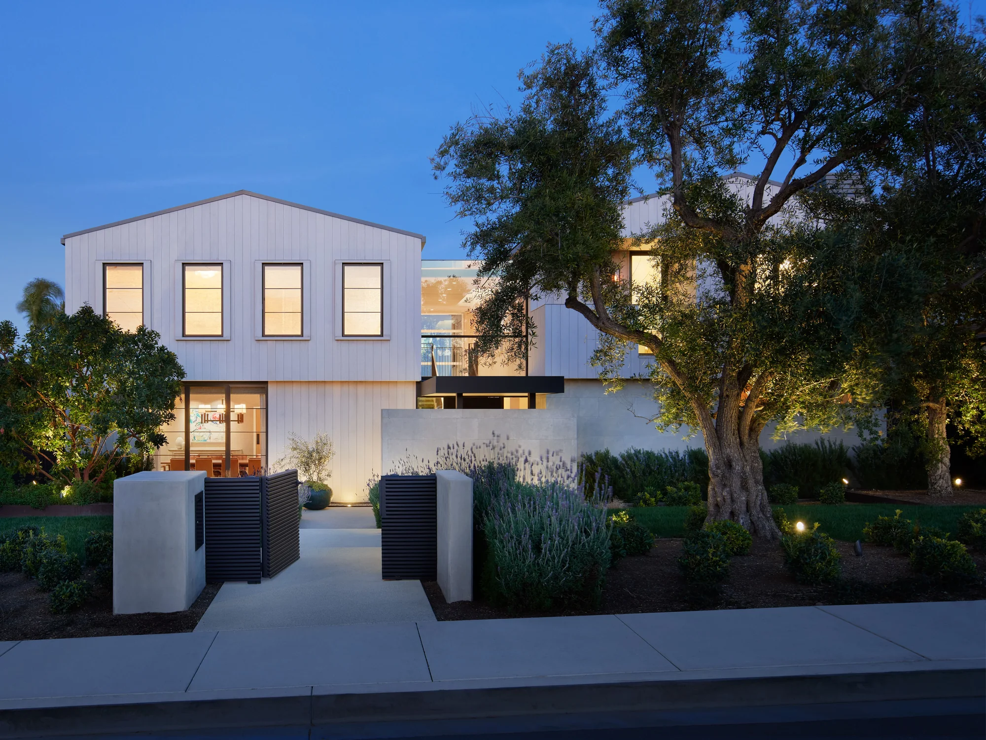 Modern white two-story home at dusk with illuminated windows, concrete entry pillars, and mature oak tree in landscaped yard.