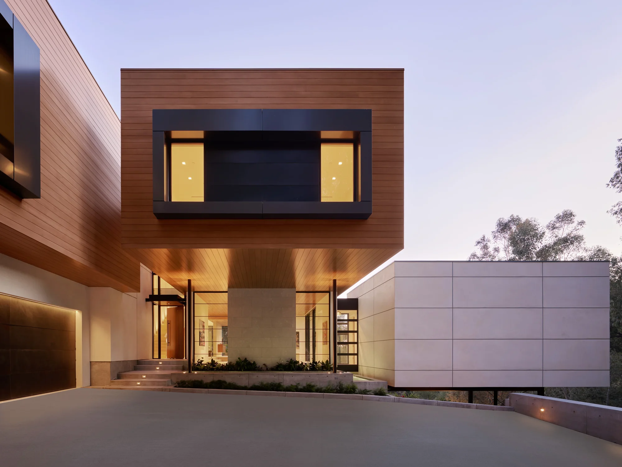 Modern custom home exterior at dusk featuring warm cedar cladding, cantilevered entry, and glowing interiors blending nature and architecture.