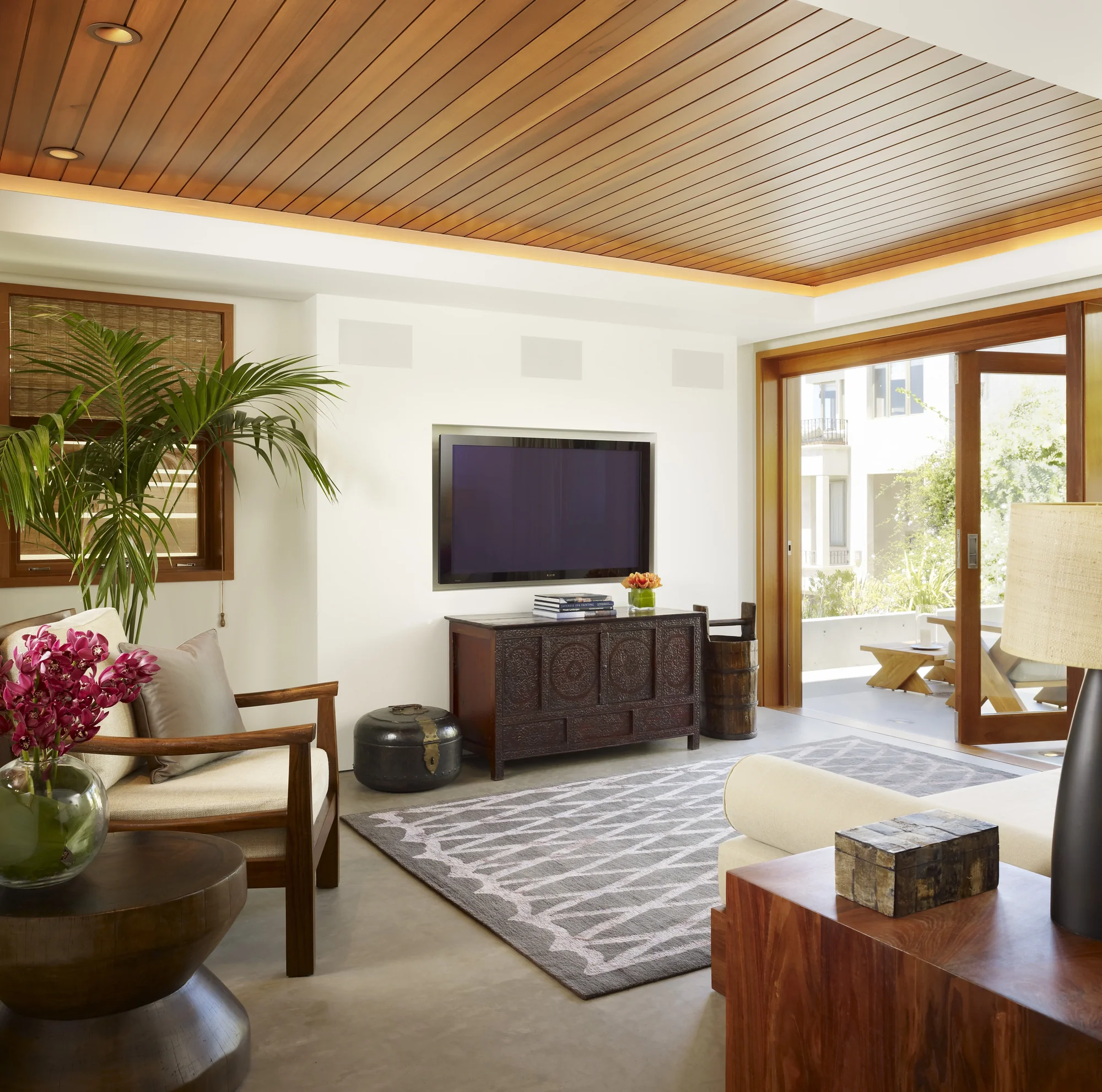 Modern living room with wood slat ceiling, TV mounted above dark wood console, opening to outdoor terrace with natural light.