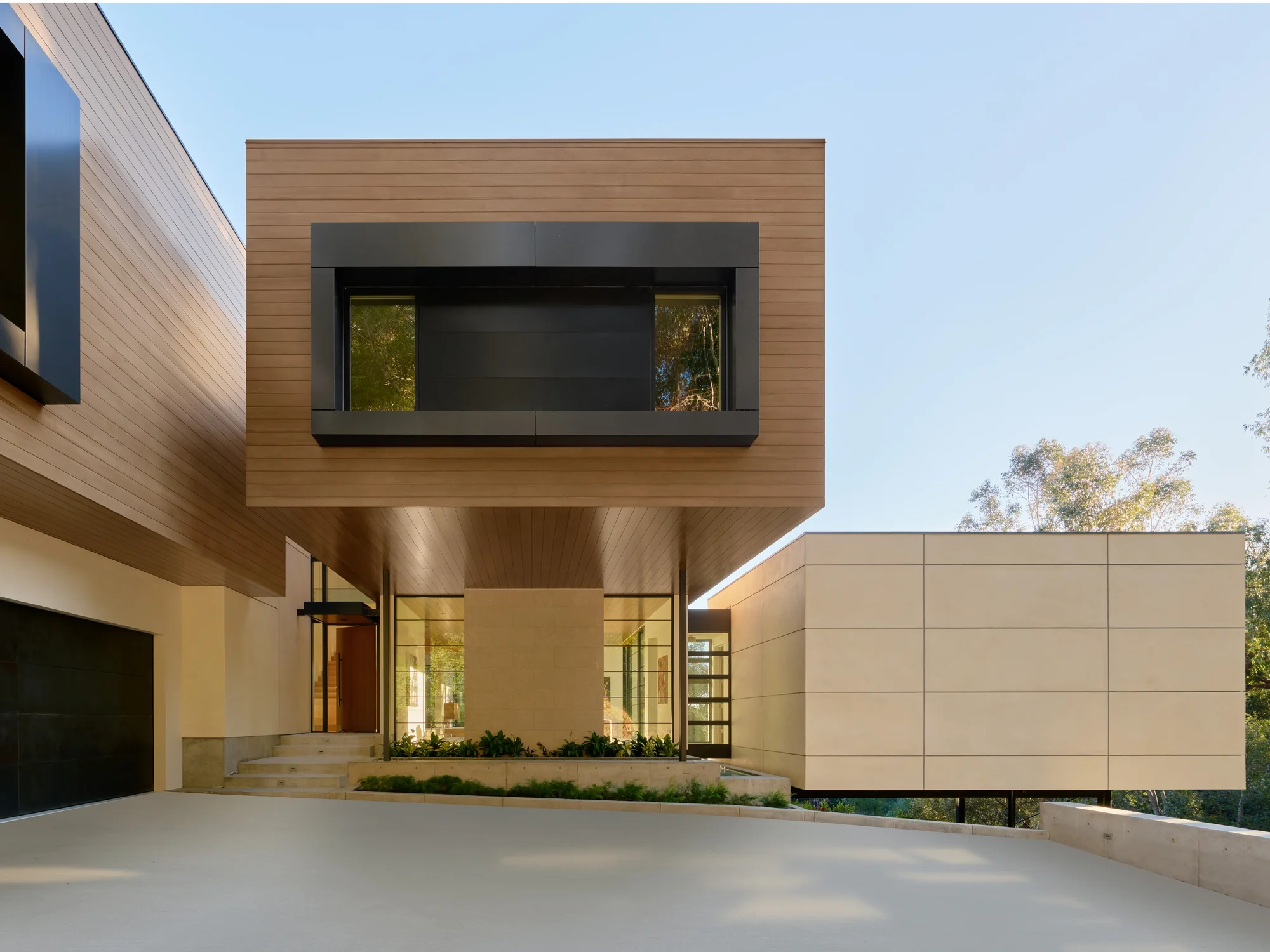 Modern home exterior featuring warm cedar cladding, dark steel windows, and a cantilevered upper volume over a sheltered entry court.