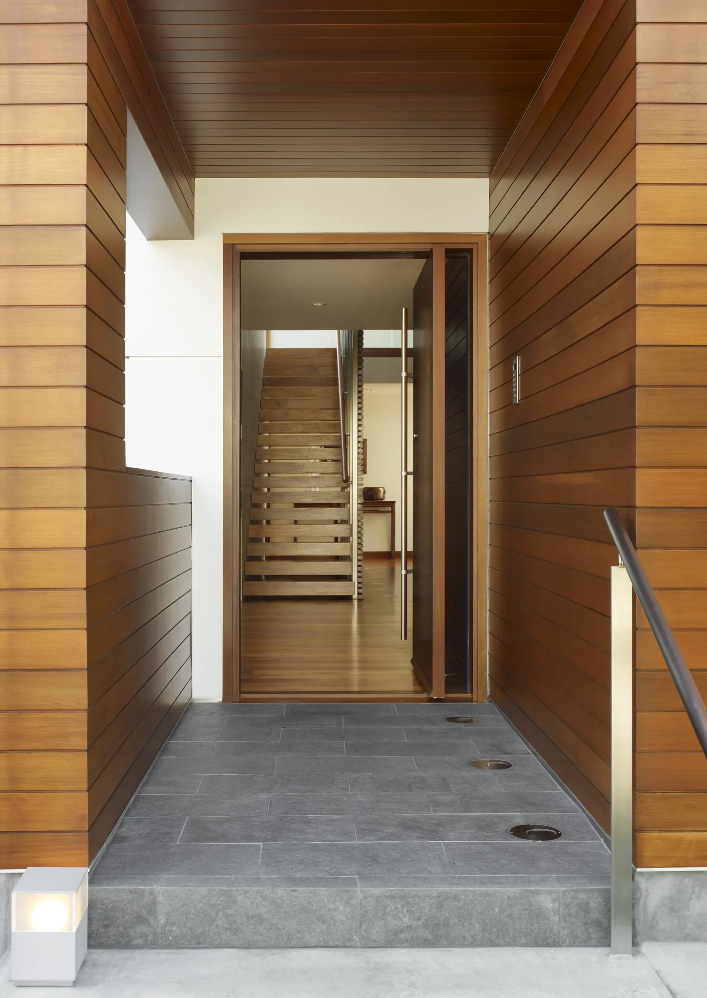 Modern home entry with glass door, warm cedar siding, white walls, gray stone floor, and view to open staircase beyond.