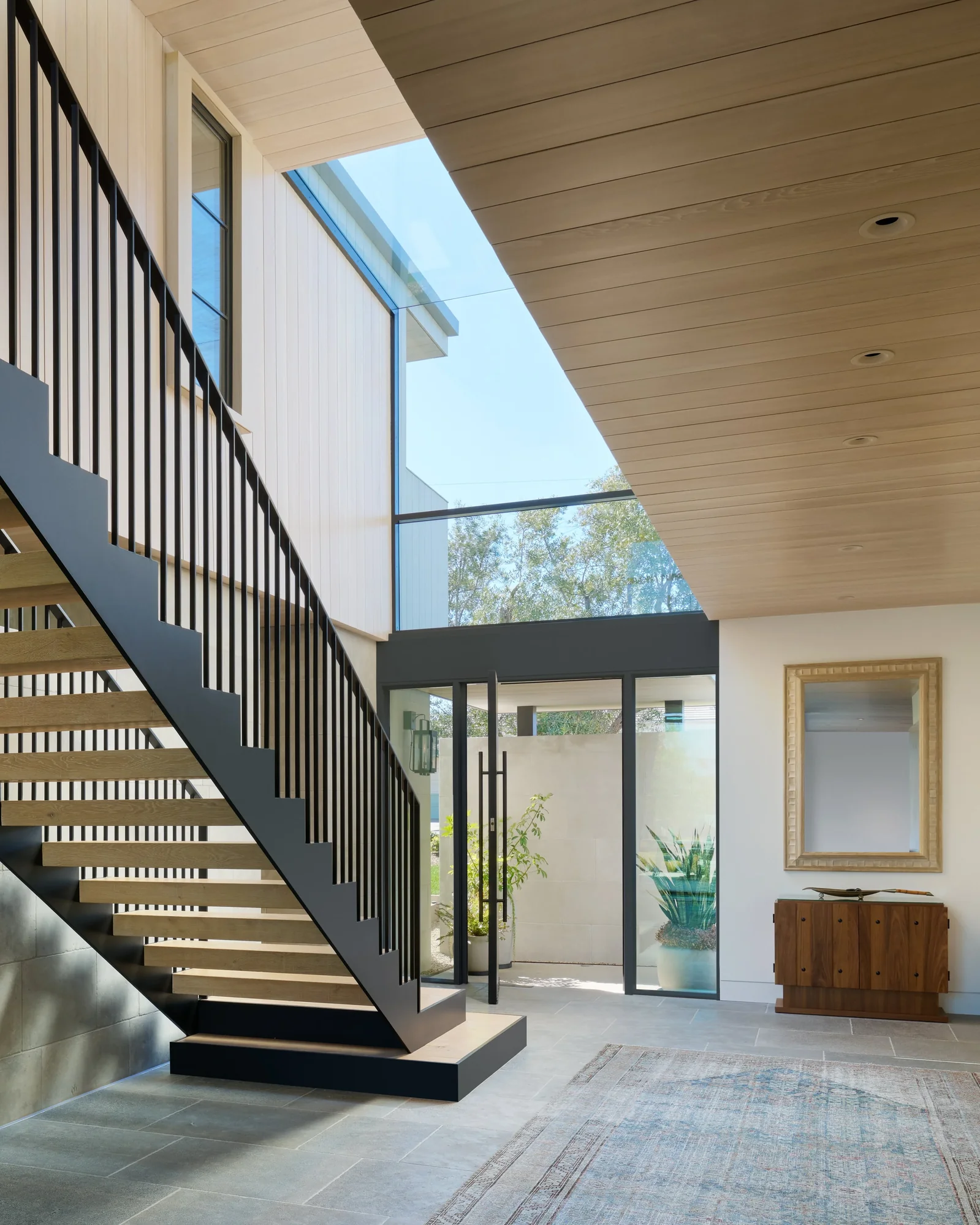 Modern entryway with black steel staircase, floor-to-ceiling glass doors, and skylight over light wood ceiling