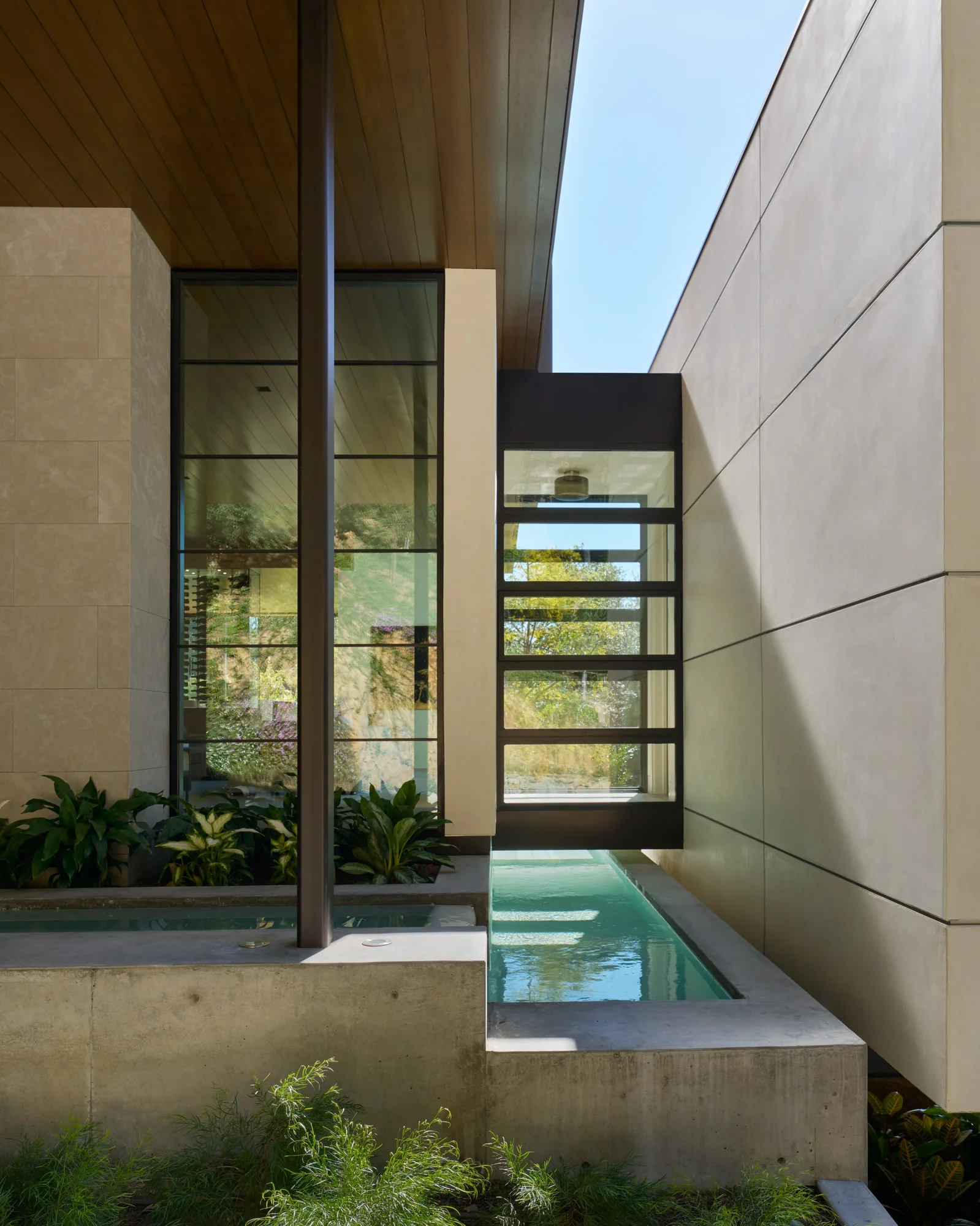 Narrow water feature leads to glass entry door framed by limestone walls and cedar ceiling, blending indoor and outdoor living.