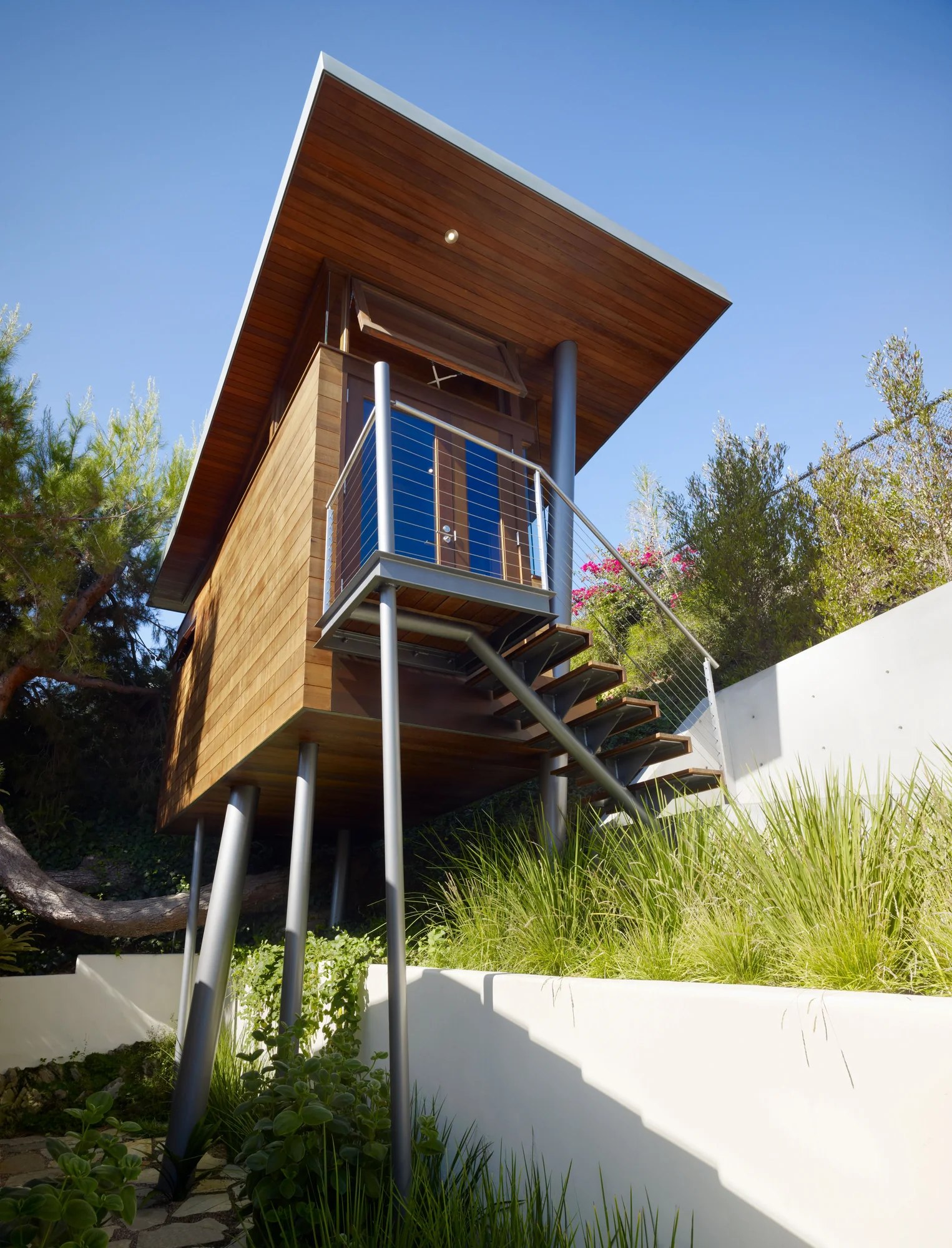 Modern cedar-clad tower structure elevated on steel columns with cantilevered roof and open stairway amid native landscaping