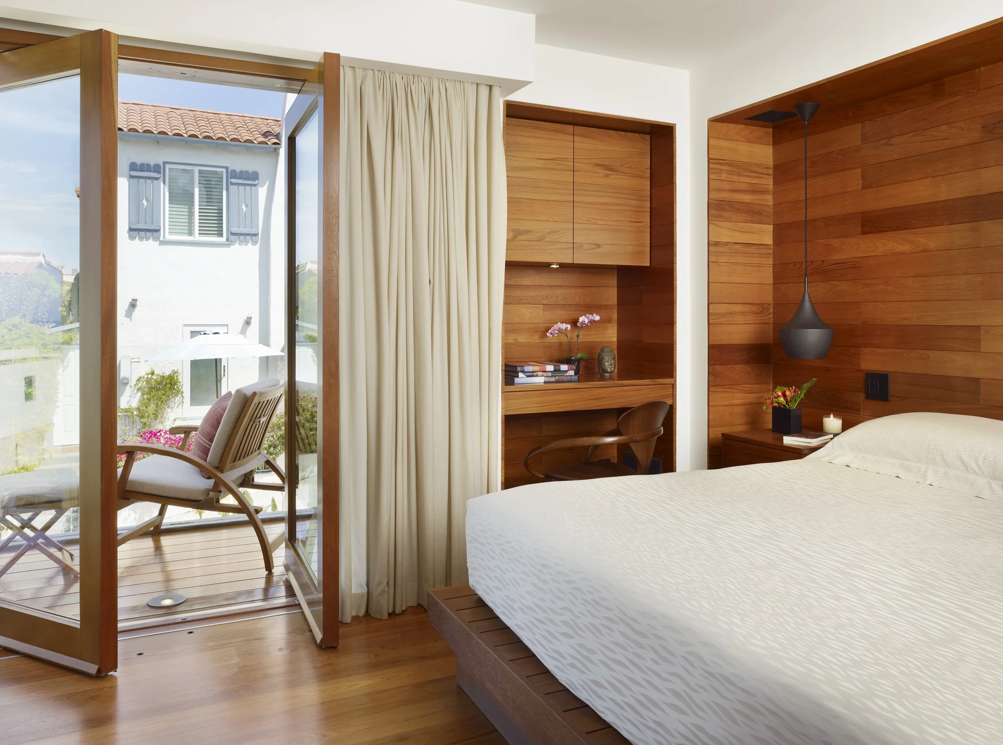 Modern bedroom with wood accent wall, built-in desk nook, and open glass door to balcony overlooking hillside homes