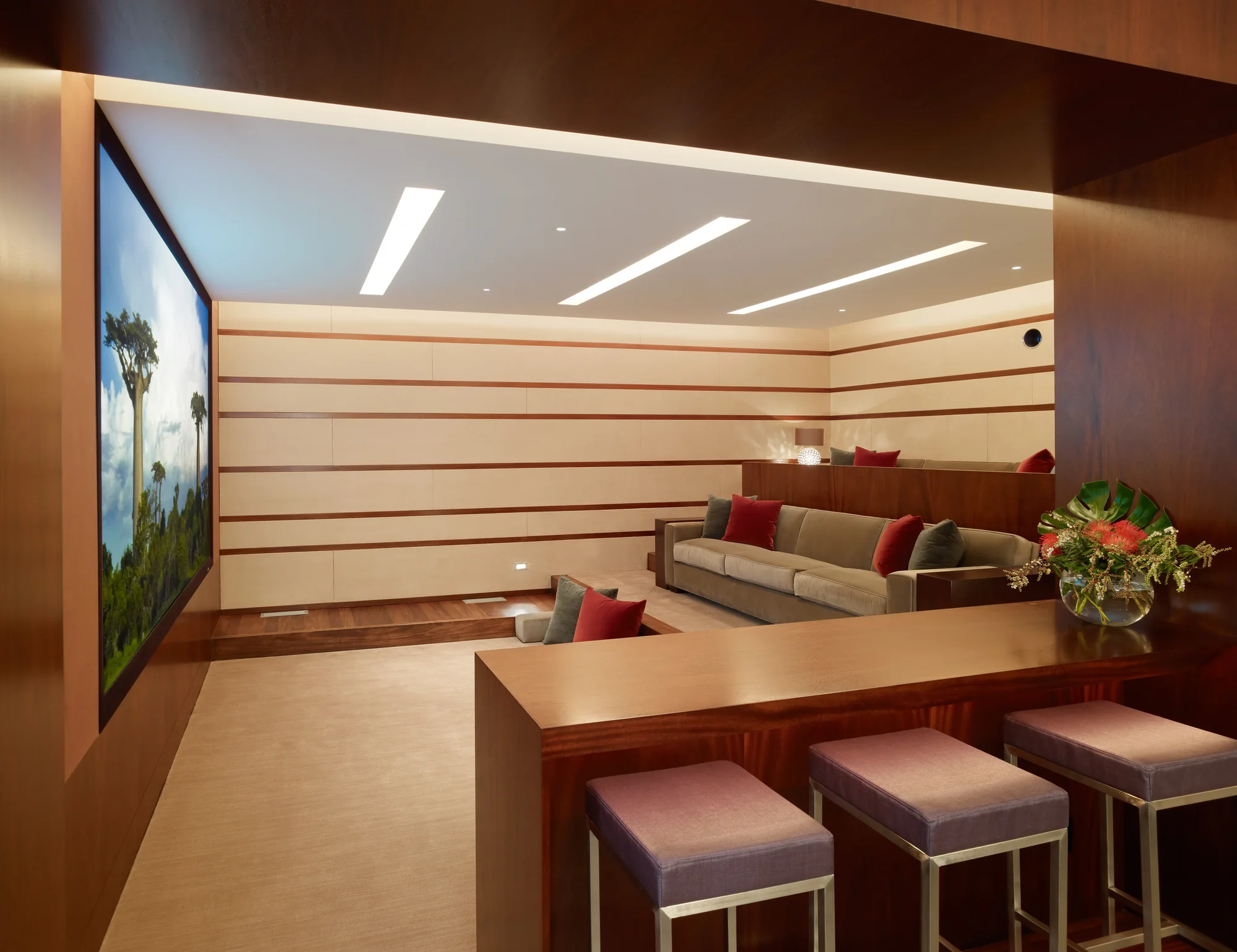 Luxury home theater with rich mahogany paneling, cream acoustic walls, red accent pillows, and a bar with velvet stools.