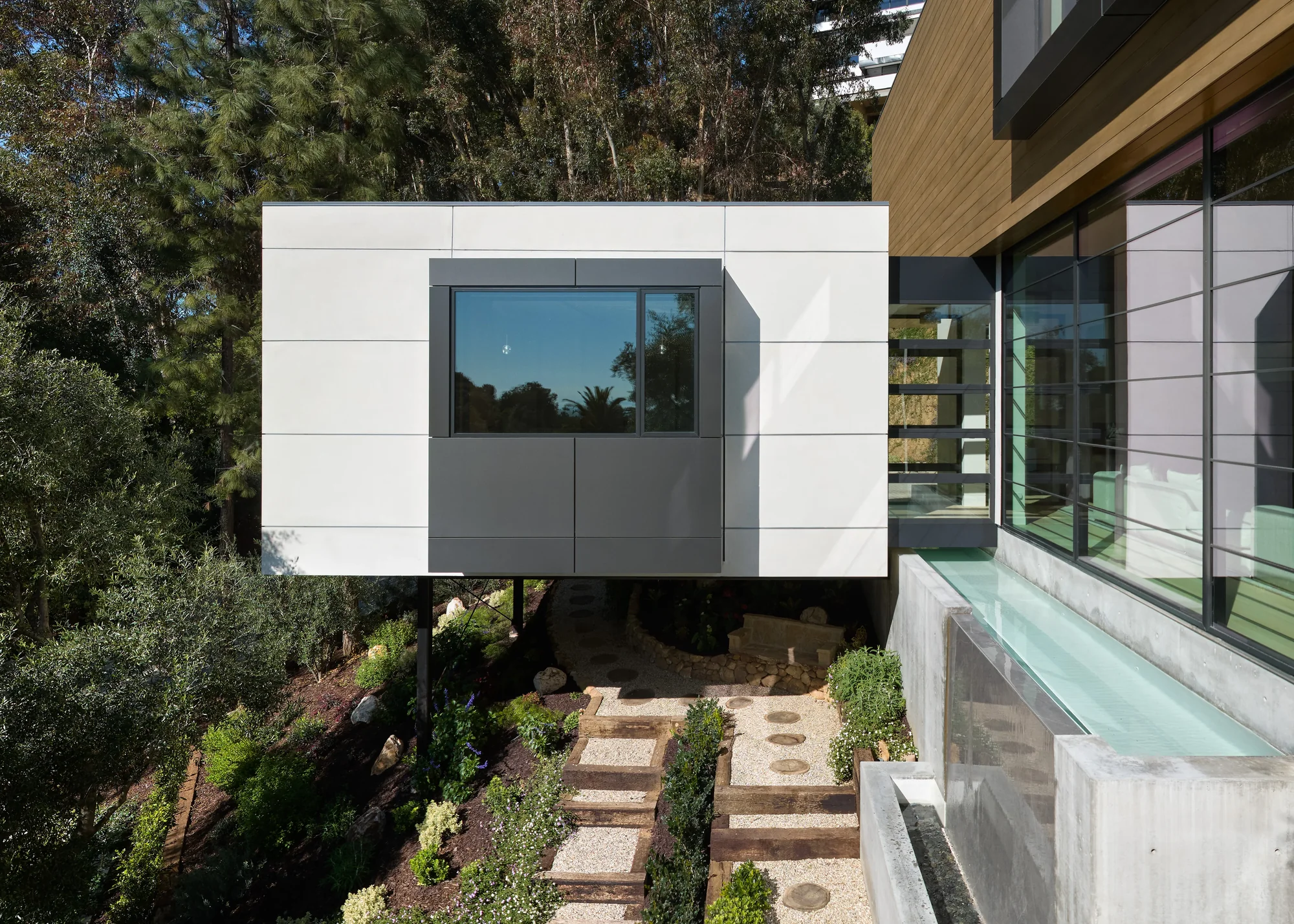 A cantilevered white panel volume hovers over a terraced garden path, connecting to a wood and glass modern residence.