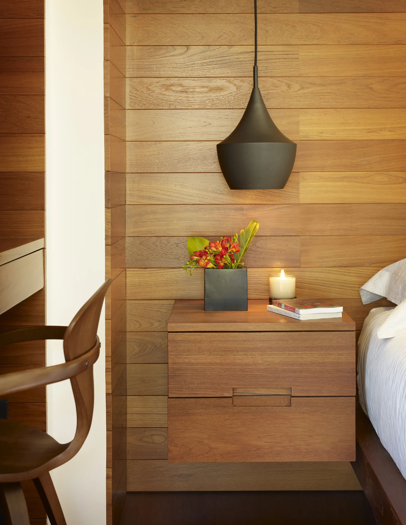 Modern bedroom nightstand in warm wood with black pendant light, tropical flowers, and candle against horizontal wood wall
