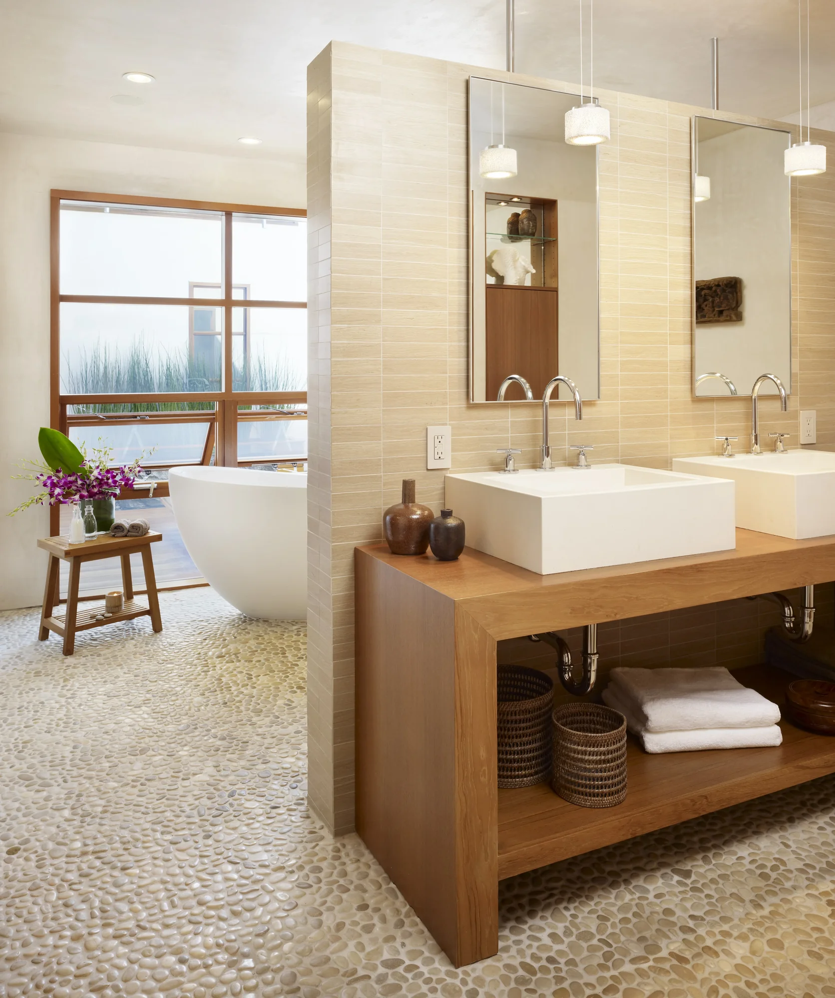 Modern bathroom with vessel sink on wood vanity, pebble flooring, and view to freestanding tub by window