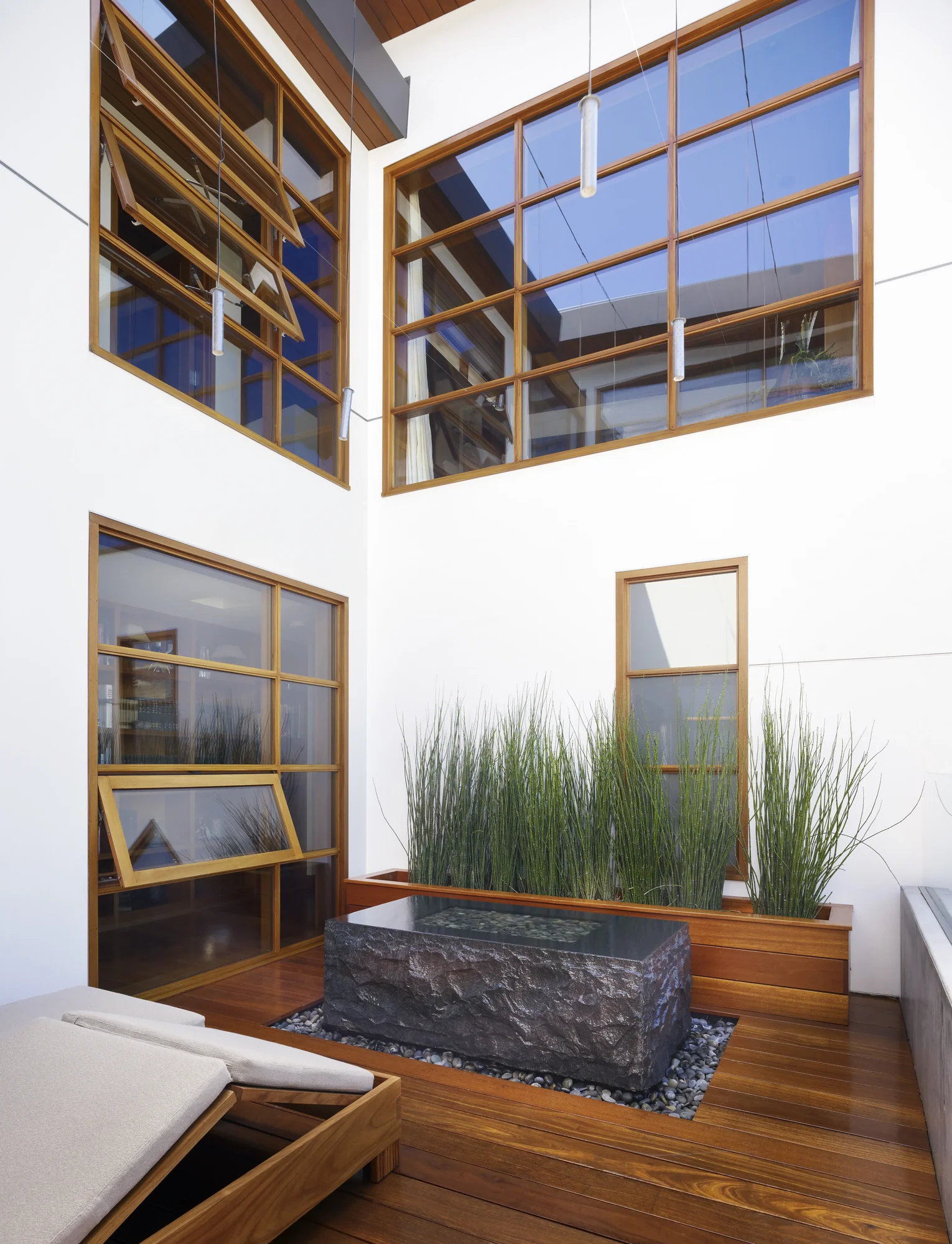 Modern interior courtyard with rough-hewn stone water feature, tall grasses, wood deck, and gridded wooden windows
