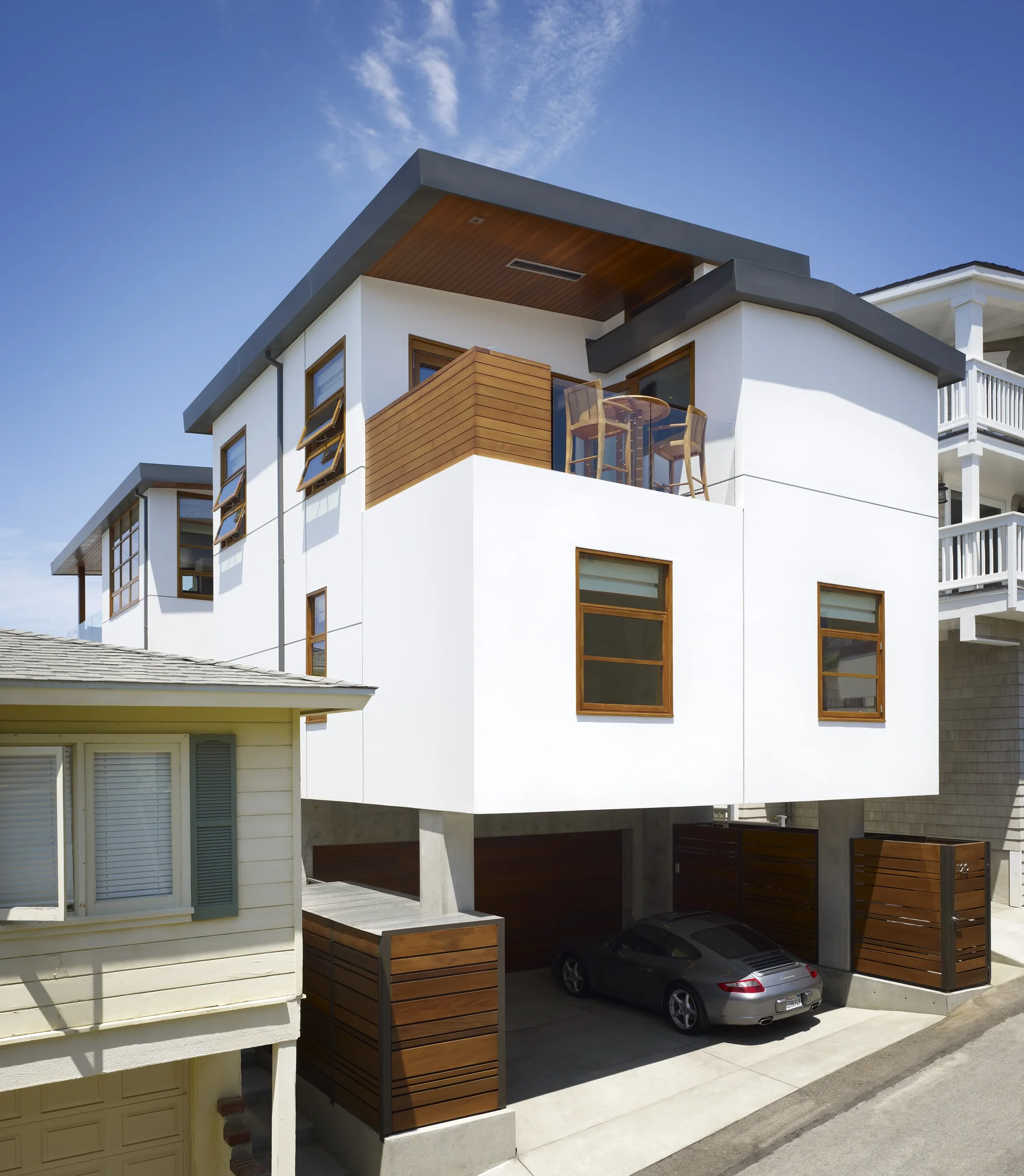 Modern three-story white stucco home with cedar accents, flat roof overhangs, and ground-level garage in coastal neighborhood.