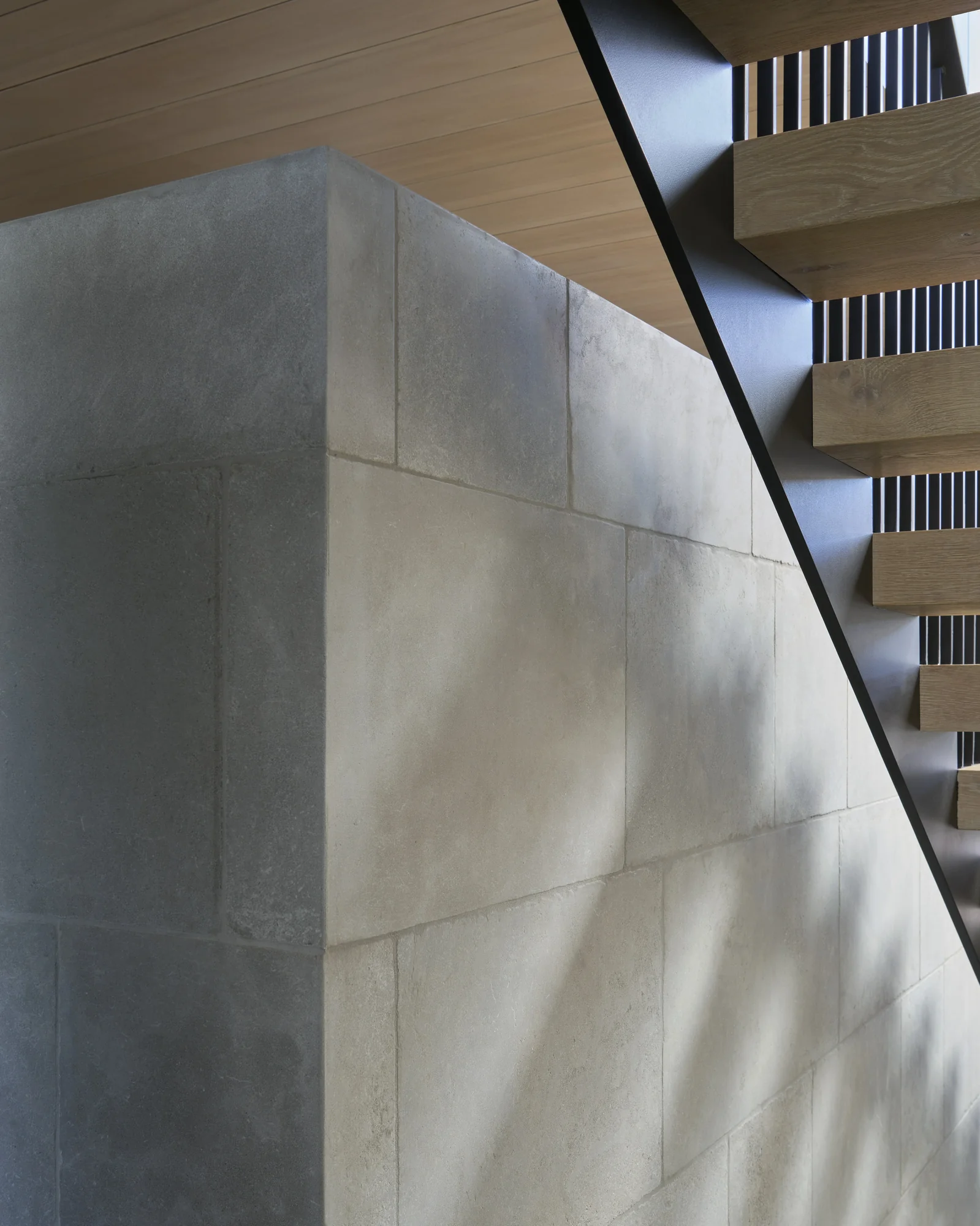 Floating wooden staircase with vertical railings beside a textured concrete wall beneath a wood-paneled ceiling