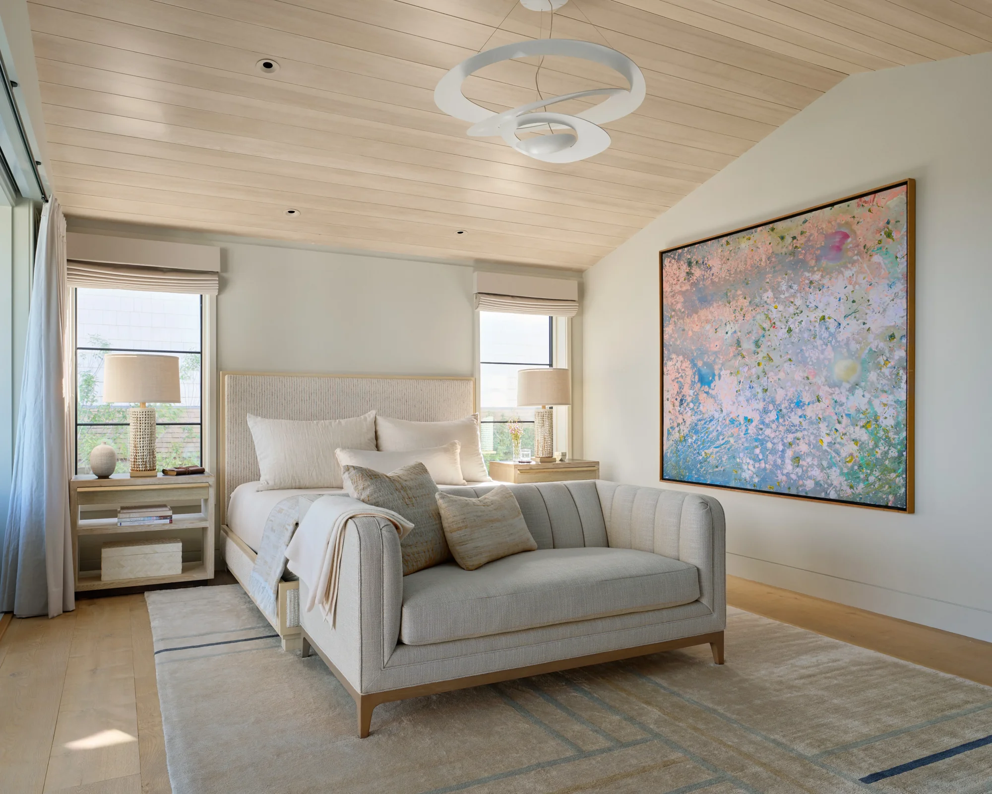 Serene coastal bedroom with light wood ceiling, abstract art, upholstered bench, and windows framing ocean views.