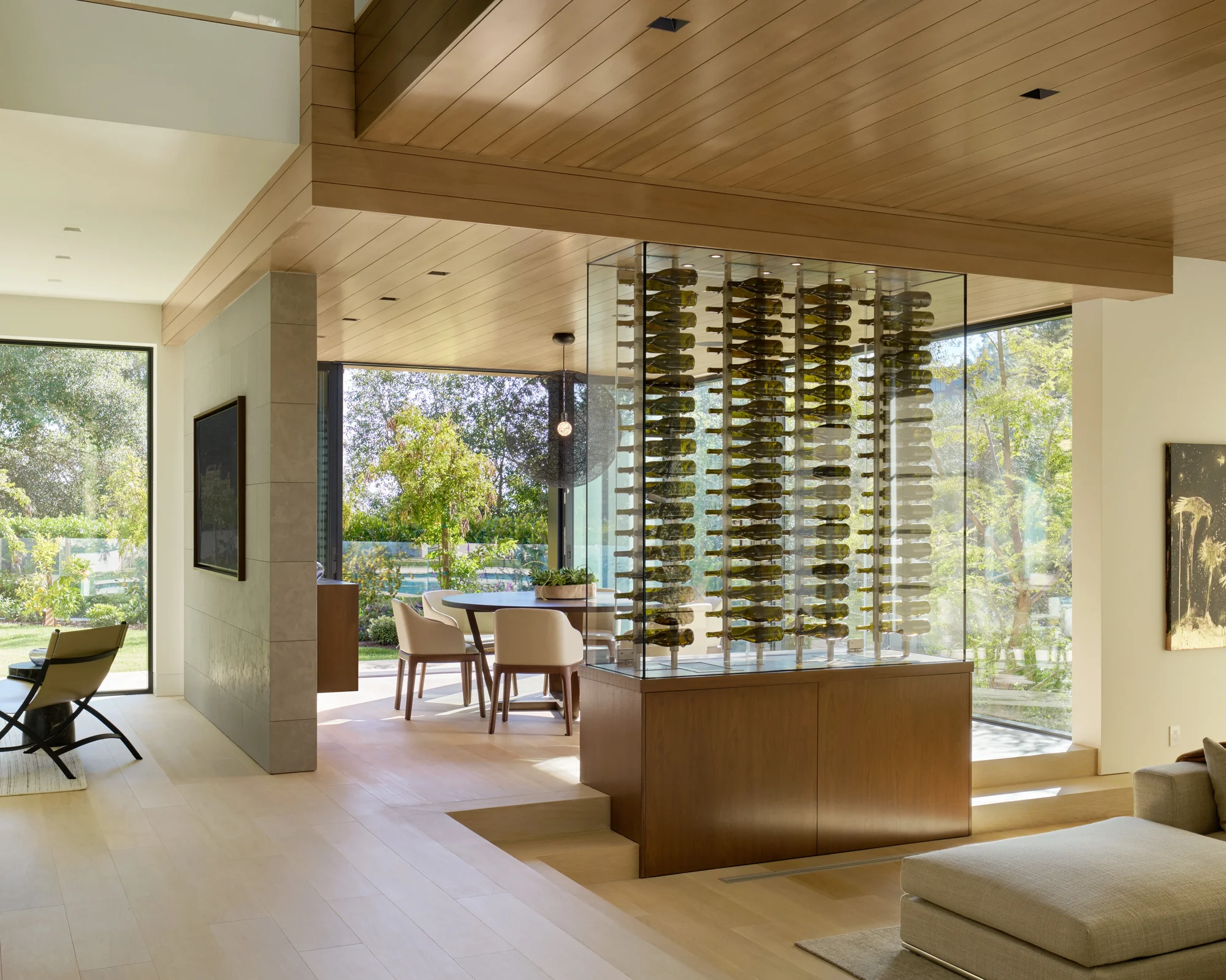 Glass wine display tower with wood base separates living and dining areas, framing lush garden views beyond.
