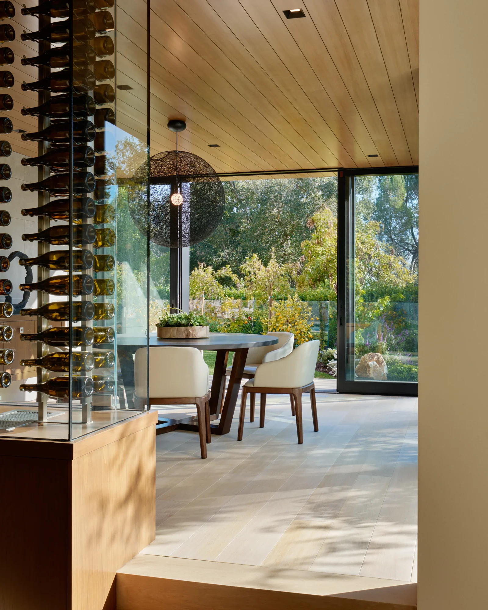 Glass-enclosed wine cellar beside a sunlit dining area with floor-to-ceiling windows overlooking a lush garden.