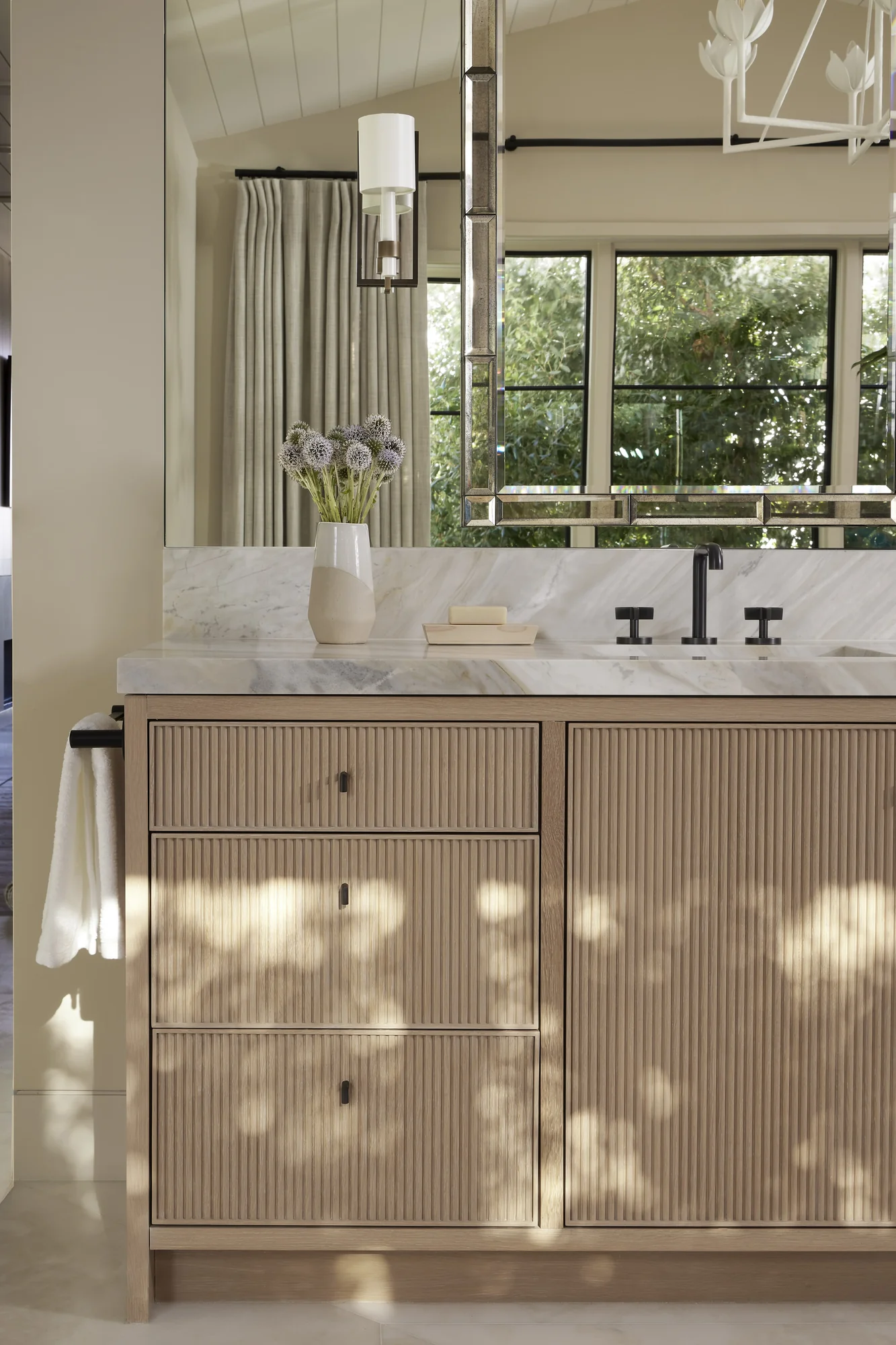 Modern bathroom vanity with fluted wood cabinetry, marble countertop, black fixtures, and window overlooking greenery.